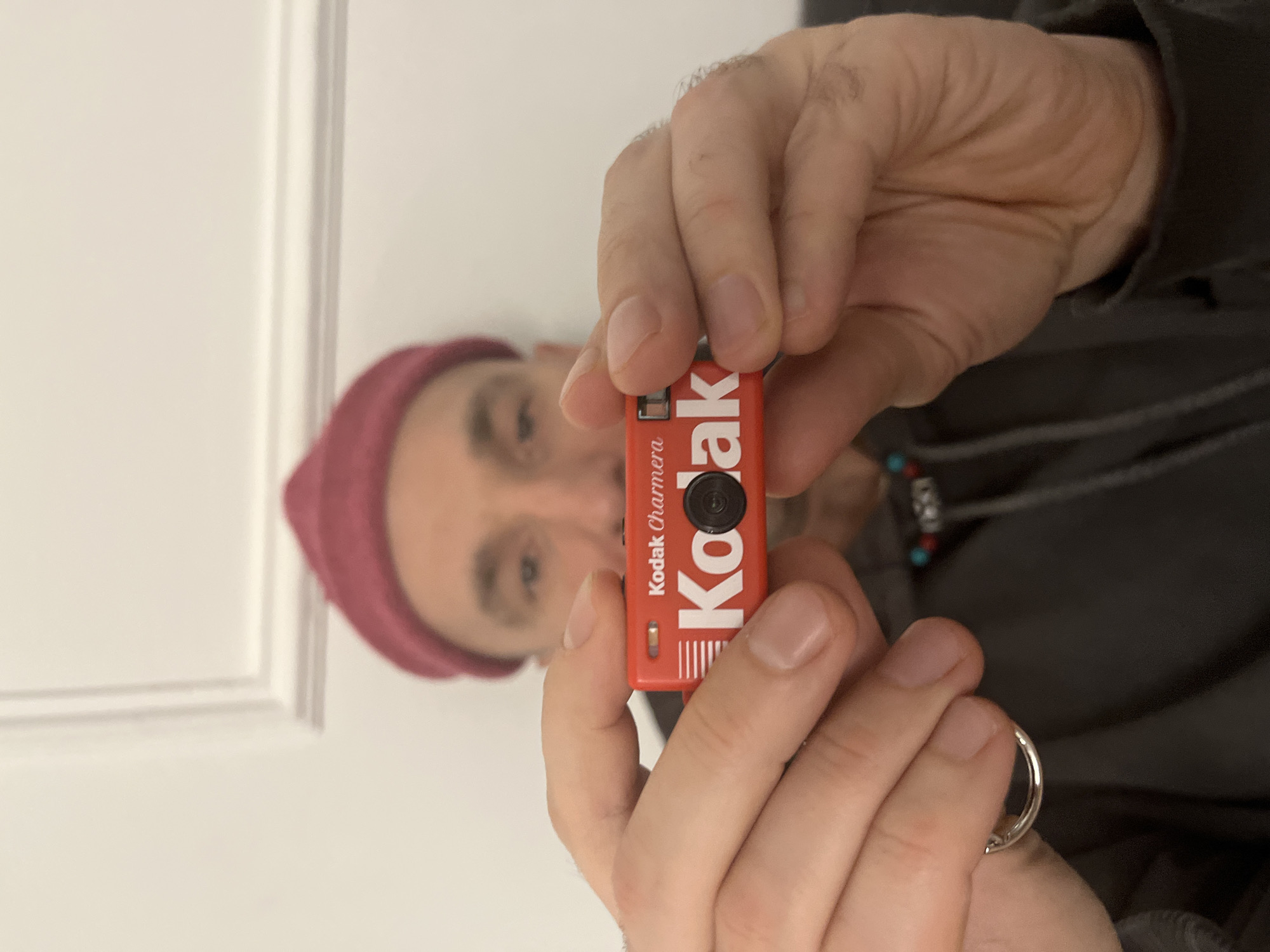 A person, partially in focus, holds up a red "Kodak Charmera" camera between the viewer and his face. The person is wearing a red beanie and a grey jacket. The camera is held in front of the person's face and is mostly in focus. The person's hands, skin tone, and fingernails are visible, as is a ring on their finger. A necklace with turquoise and red beads is partially visible. The background is a white wall and a partially visible white door frame. The location in the city of Budva, Montenegro is not possible to determine from the image.