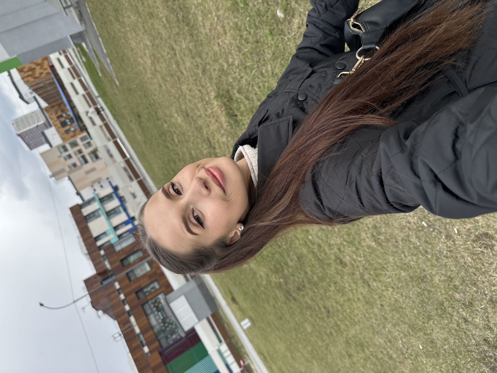 The photograph shows a woman with long brown hair lying on a grassy field. She is wearing a black quilted jacket and a light-colored sweater. Her hair is spread out on the grass near her. In the background, there are modern buildings with multi-colored facades. The location is Yekaterinburg, Russia.