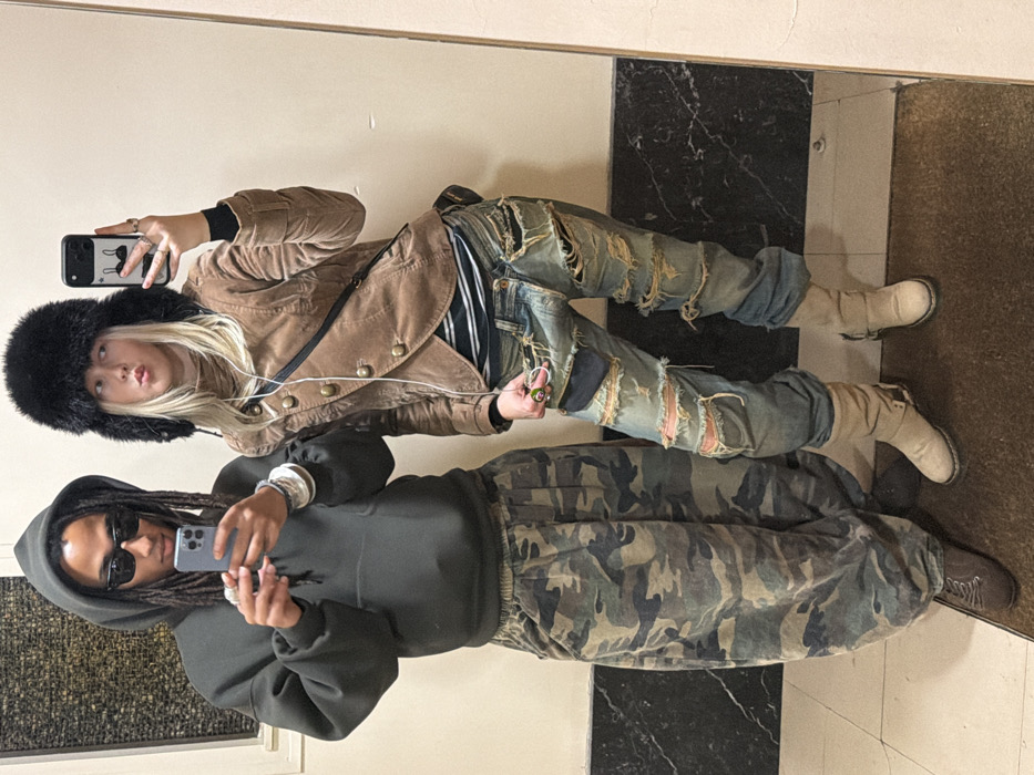 Two individuals are captured in a mirror selfie, standing upright against a light-colored wall with a tiled floor visible below them.
On the left, a person with blonde hair is wearing a large, fluffy black hat that covers their ears. They have light skin and their lips are pursed in a slight pout. They are wearing a tan or light brown jacket that appears to be made of velvet or corduroy, featuring brass-colored buttons down the front and on the cuffs. Underneath, a striped top in dark and light colors is visible. Their jeans are light wash, heavily distressed with large rips and tears, revealing patches underneath. On their feet are light-colored, possibly suede or nubuck, boots with a chunky sole and some buckling detail around the ankle. They wear several rings on their fingers, including one with a prominent green stone. A black shoulder bag strap crosses their body. They are holding a black smartphone with a white case, which features a simple black outline drawing (resembling a stylized animal or abstract shape) on the back, and are looking directly into the phone's camera, presumably taking the picture. Earphone wires are visible hanging from their ears.
On the right, a second person with dark skin and dreadlocks is wearing dark sunglasses. They are dressed in a dark grey or charcoal-colored oversized hoodie with gathered cuffs and hem. Their pants are a camouflage pattern in shades of green, brown, and black. They are holding a silver or grey smartphone in their hands, with some rings and bracelets visible on their wrists. Their footwear appears to be dark-colored shoes or boots.
The setting appears to be an indoor space. The floor visible in the reflection features large square tiles, with one section being a dark, possibly black, marble-like pattern and an adjacent section being a lighter, plain brown or tan color. The wall reflected behind them is plain and light-colored. The overall impression is a casual, contemporary portrait of two people showcasing their fashion.