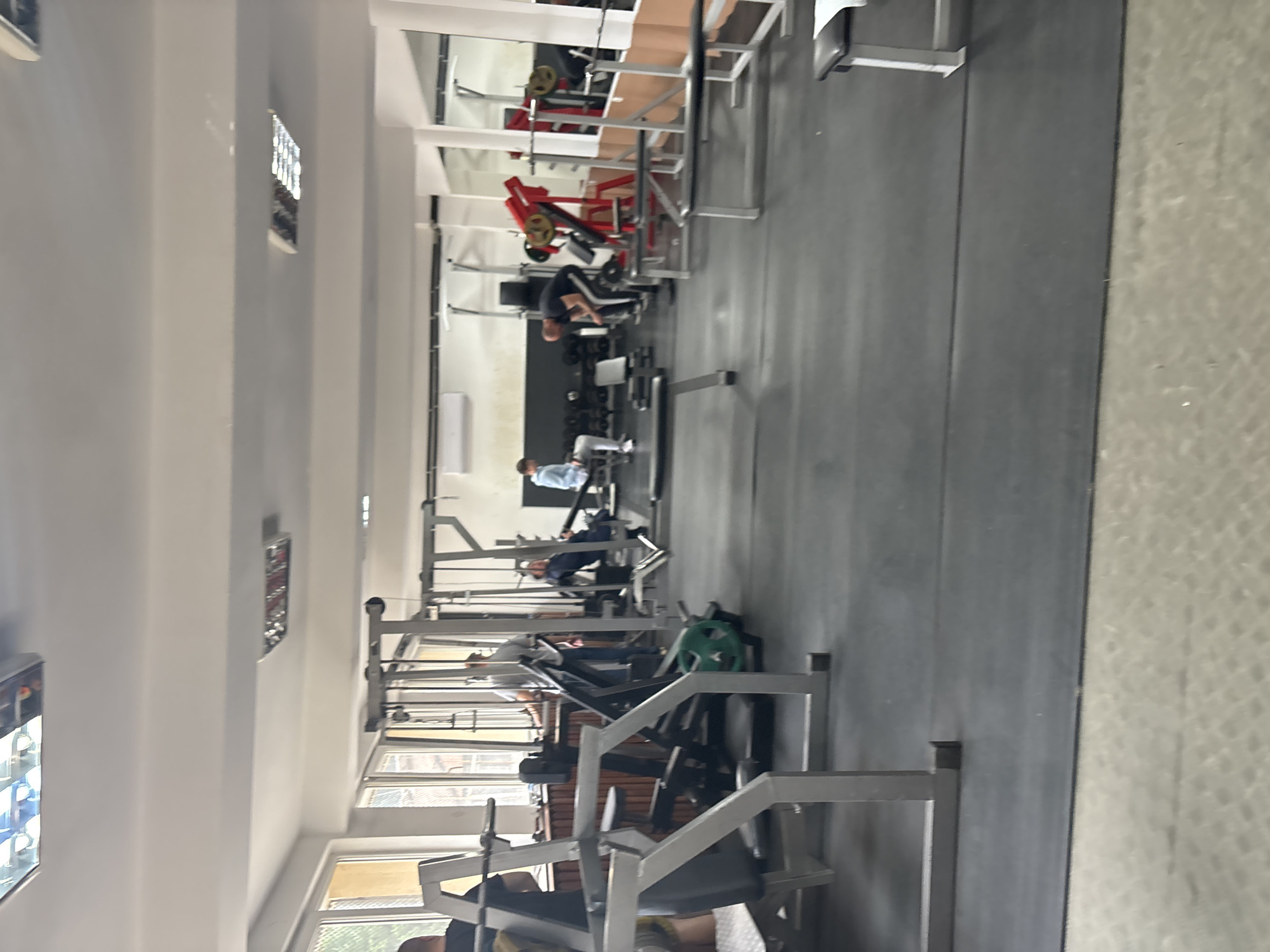 This media file captures an indoor view of a gym, likely in Zlatograd, Bulgaria, as per the provided context. The scene is oriented vertically in the original image but depicts a standard horizontal layout for a gym.
**Subjects:** Several individuals are present, engaged in various exercises or resting. In the foreground, a person in dark attire is seated on a weight training machine, facing away from the viewer. Further back, another person wearing a blue top is using a machine, possibly a leg press or similar, also facing away. Other individuals are visible in the mid-ground and background, interacting with equipment.
**Objects:** The gym is equipped with numerous strength training machines, including those with red and grey frames. Various weight plates, some green and some yellow, are loaded onto machines or stored on racks. Dumbbells are partially visible on a rack in the middle ground. The floor is covered with dark, possibly black, rubber matting typical of a gym. The walls and ceiling are white, and fluorescent lighting fixtures are visible on the ceiling. Windows along one side of the room provide natural light. Mirrors on the far wall reflect parts of the gym interior.
**Activity or event taking place:** The people in the image are actively exercising or preparing for workouts, indicating a typical gym environment.
**Notable details:** The gym appears well-lit, benefiting from both natural light entering through windows and artificial overhead lighting. The time of day appears to be daytime, judging by the ambient light. No specific weather conditions are discernible from the interior. There is no visible text or branding on any equipment or surfaces within the image. The overall impression is one of a functional and active fitness space.