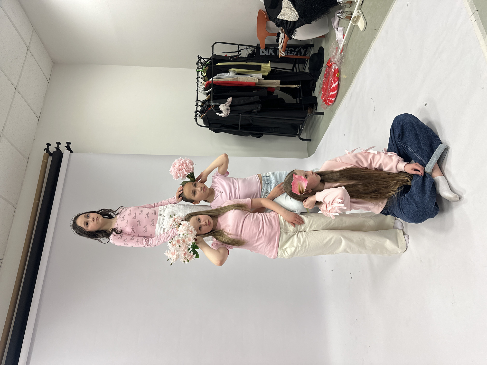 The image depicts four girls lying on a white backdrop in what appears to be a studio setting. The girl on the left wears a pink long-sleeved shirt with "Barbie" written on it, paired with white pants and a tiara. The next girl wears a pink t-shirt with white pants. The third girl also wears a pink t-shirt, paired with denim shorts. The girl on the right wears a light pink long-sleeved shirt with bows on the sleeves, paired with blue jeans. Each girl holds pink flowers. A clothing rack filled with clothes, shoes, and accessories, including a "Birthday" sign, is behind them. The location of the city of Votkinsk, Russia is unknown.