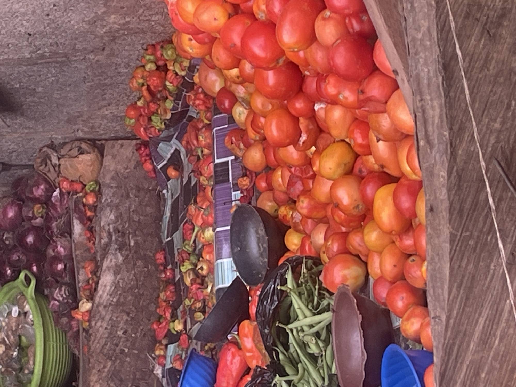 The image depicts a vendor's stall in Abuja, Nigeria. There are piles of red tomatoes, onions, peppers, and green beans for sale. Some items are arranged on a checkered fabric. The stall is constructed of wood and cement.