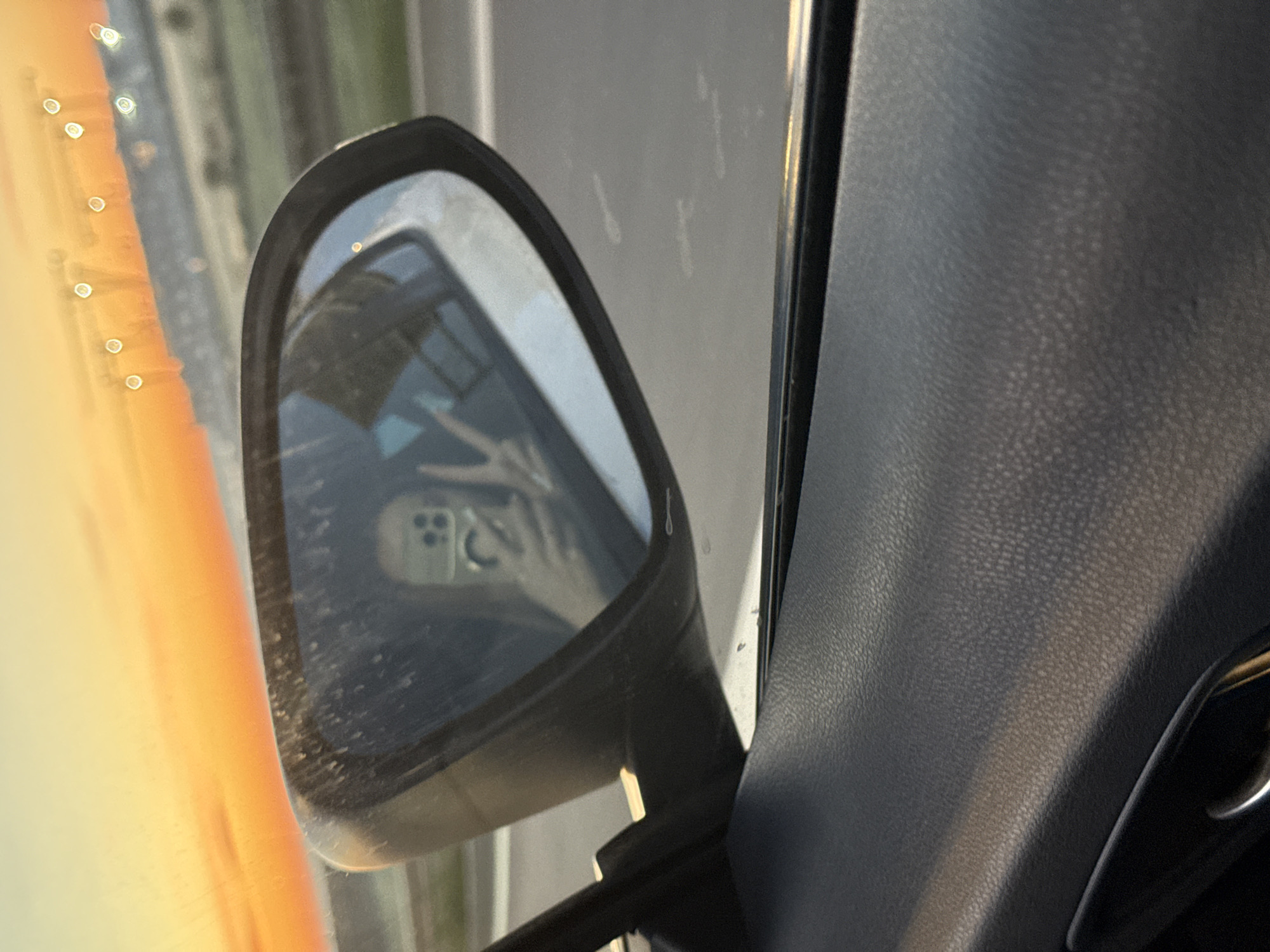 A photograph taken from inside a vehicle shows a side mirror reflecting a hand displaying a peace sign and a smartphone. The setting appears to be a roadway during sunset, characterized by the orange hue visible. The interior of the vehicle is visible in the frame, including a portion of the textured dashboard. The precise location within Surcin Urban Municipality, Serbia cannot be determined from this image alone.