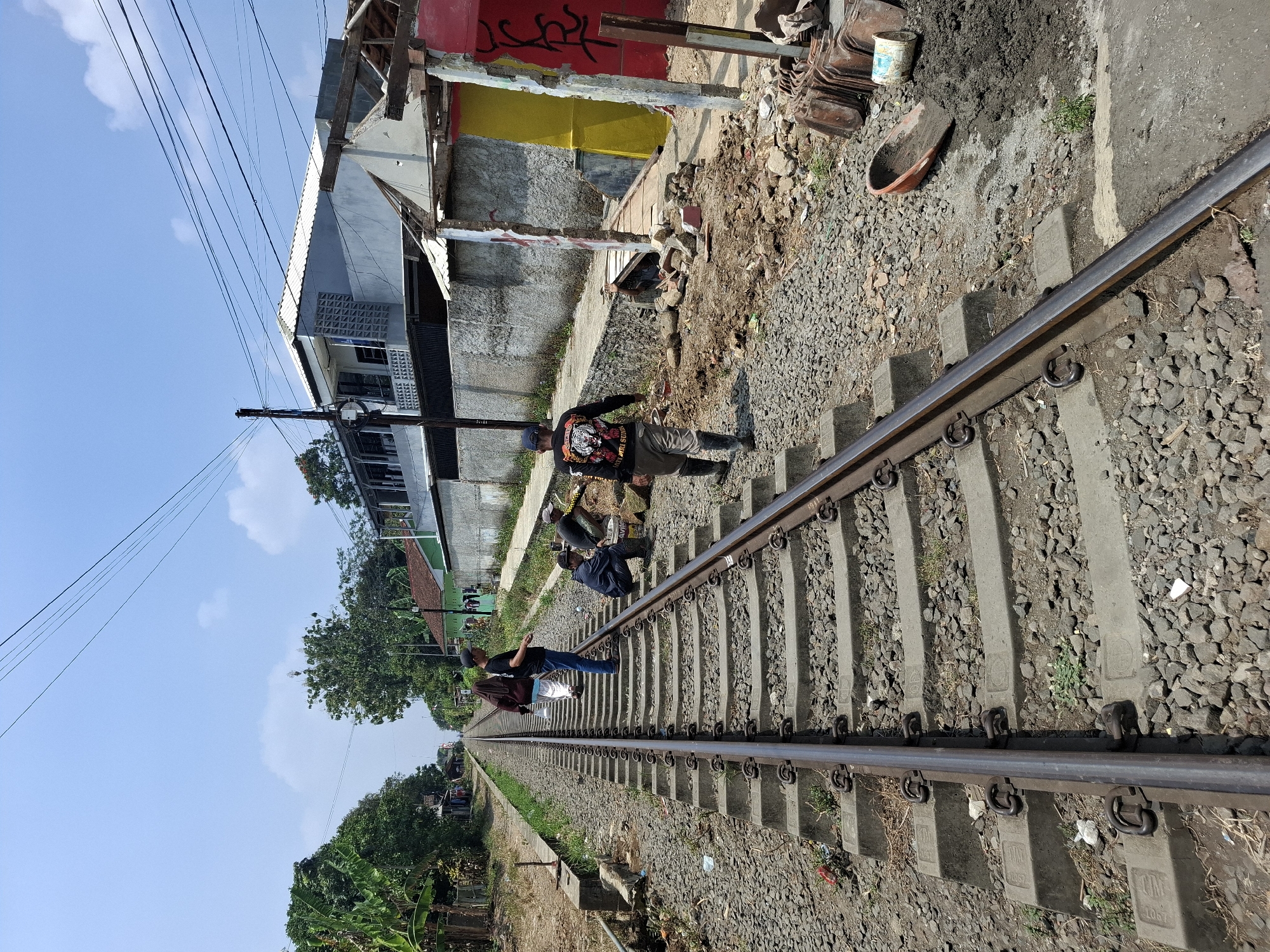In Karangtengah, Indonesia, a group of individuals are gathered near a set of railroad tracks. One person, wearing a black long-sleeved shirt with a tiger graphic and a black cap, is walking away from the camera and towards the right side of the image. Another individual, in a blue jacket and black pants, is crouching near a white bucket. A third person, in a dark blue jacket, is also crouching and appears to be working. Two other individuals are walking away from the camera on the train tracks, one in a brown hijab and the other in a white outfit. The scene is set under a clear blue sky with scattered clouds. The railroad tracks are surrounded by gravel and concrete sleepers. To the right of the tracks, there is a building with a red and yellow facade, appearing partially constructed or in disrepair, with debris and building materials scattered around. Further in the background, more buildings and trees are visible.