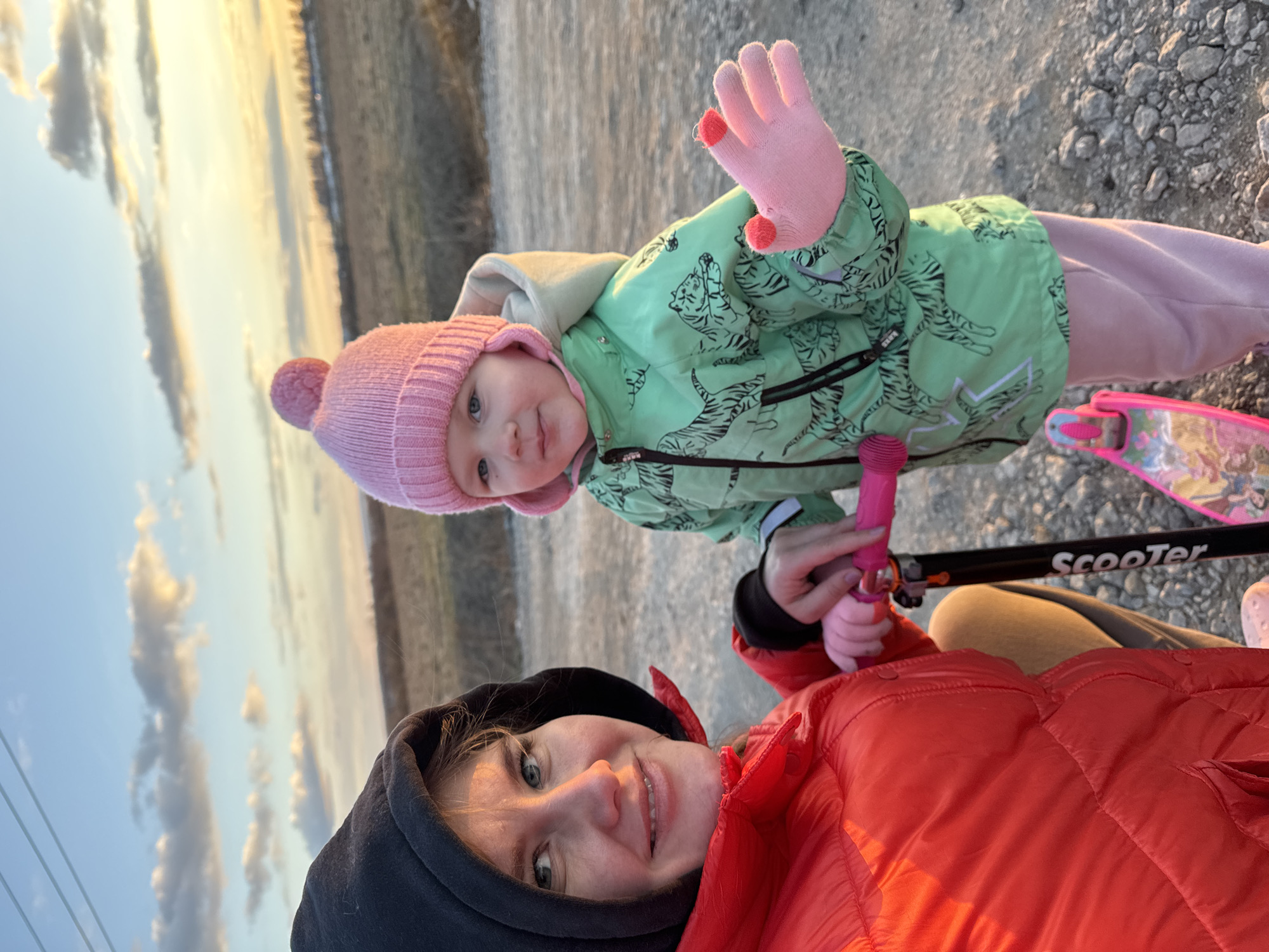 A candid, selfie-style photograph captures an adult woman and a young child outdoors during the golden hour of late afternoon or early evening. The woman, positioned in the foreground on the right, smiles warmly at the camera. She has light hair and blue eyes, and wears a red puffer jacket over a black hooded sweatshirt.
To her left and slightly behind her, a young child, likely a girl, also looks towards the camera with a gentle, curious expression. The child is dressed in warm clothing, including a light pink knitted beanie with a pom-pom, a light green jacket adorned with black tiger patterns, and pink pants. Her hands are covered in pink mittens with distinct red fingertips. She is holding onto the handlebars of a pink scooter, on which the word "Scooter" is visibly printed on the frame.
The setting appears to be a gravel or rocky path. In the background, the sky displays warm, golden and light blue hues with scattered clouds, indicative of the low sun. An open, natural landscape, possibly a field or distant trees, stretches towards the horizon. Both individuals appear content and happy in the outdoor environment.