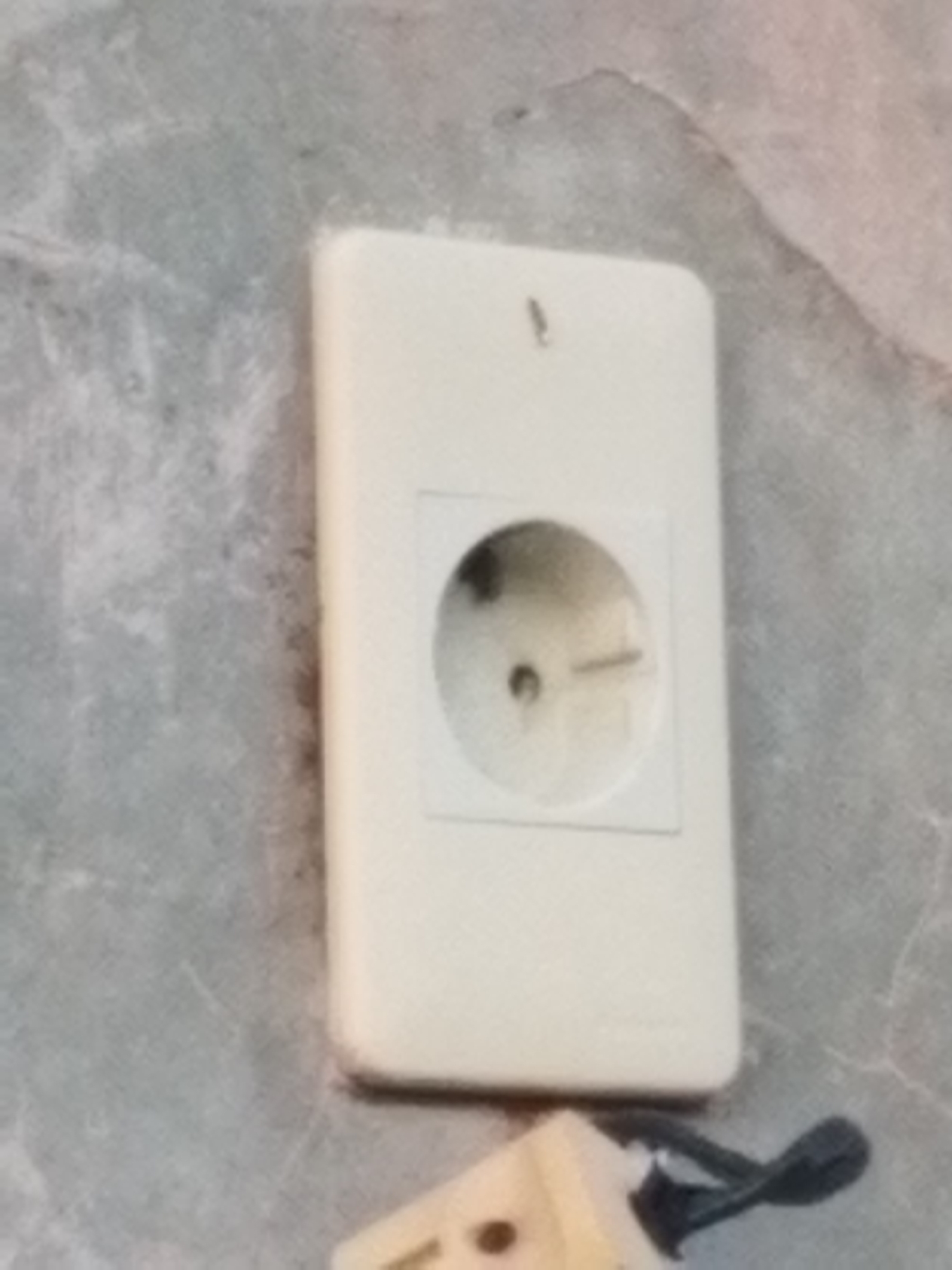 A close-up shot shows a European-style electrical outlet mounted on a marbled-looking wall. The outlet is white and rectangular, with a circular recessed area in the center containing the plug receptacle. A two-pronged plug is partially visible at the bottom of the frame, with a black cord extending away. The location appears to be indoors. There is no information about the location being in the city of Lhokseumawe, Indonesia. No people are visible in the image.
