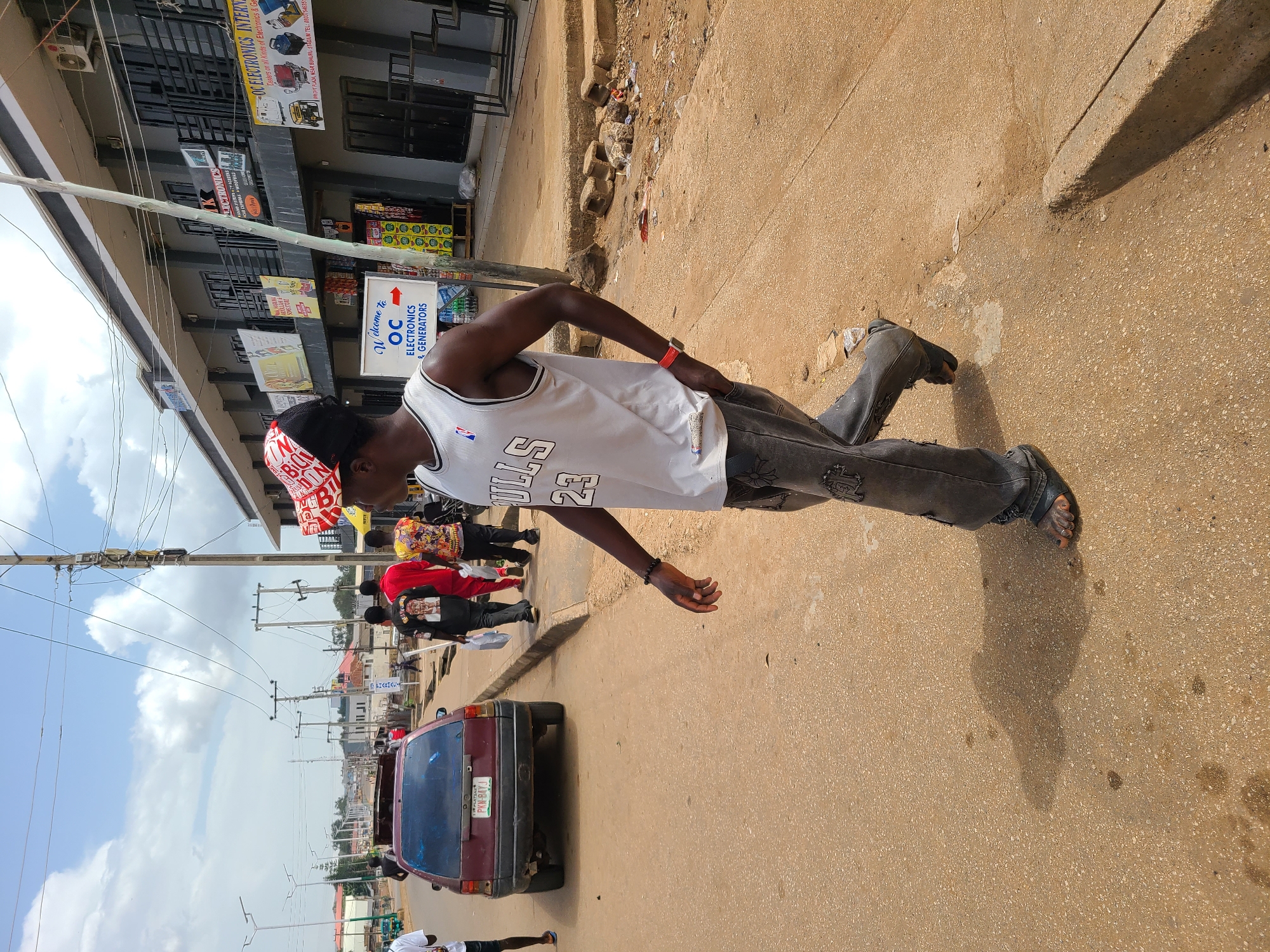 The image captures a street scene in an urban area of Unknown, Unknown. A man wearing a red and white cap, a white Bulls jersey with the number 23, black jeans, and sandals is walking on the sidewalk. Behind him, there's a red car with license plate PKN-B4YJ, and a group of people walking on the street. The storefronts are adorned with signs advertising electronics and pharmacies. Overhead, power lines stretch across the sky, which is partly cloudy. The sidewalk is concrete with visible wear and discoloration.