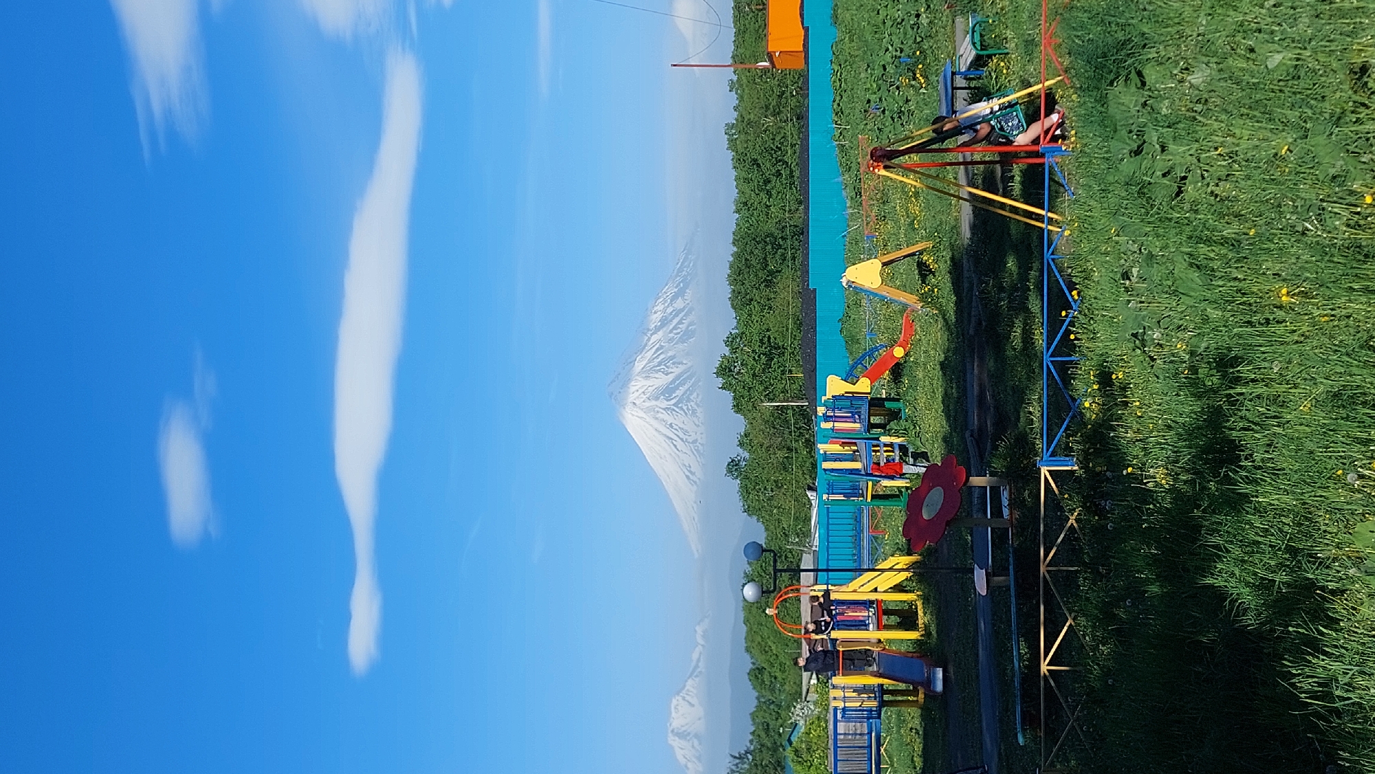 This image captures a vibrant outdoor scene with a playground and a majestic snow-capped mountain in the background. The playground features a variety of colorful structures, including slides, swings, and climbing frames, designed for children's recreation. A few people are visible on the playground, engaging in activities or resting. The setting appears to be a park or recreational area, with lush green grass and scattered yellow flowers surrounding the playground. The mountain, possibly a volcano, dominates the horizon, its snowy peak contrasting with the clear blue sky and scattered white clouds. The overall atmosphere suggests a pleasant day, likely during daylight hours. The image's orientation is rotated, with the sky and mountain appearing on the left side and the playground and grassy area on the right. No text is visible in the image.