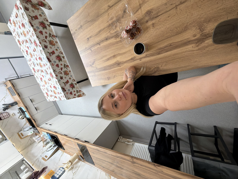The image displays a modern kitchen and dining area located in the city of Sarapul, Russia. A blonde woman is positioned in the foreground, taking a selfie with her right arm extended towards the camera. She wears a black short-sleeved top, and tattoos are visible on her left arm. Her left hand is resting near her neck. She is situated at a light-brown wooden table, on which a dark-colored mug and a clear plastic container holding approximately six frosted pastries are present. The kitchen in the background features white base cabinets with dark handles, a wooden countertop, a black sink, a black built-in cooktop, and a white marble-patterned backsplash. A stainless steel refrigerator is partially visible. Another dining table, covered with a festive tablecloth depicting reindeer, bells, and other holiday motifs, is further in the background. The flooring is light gray, and a black metal shelving unit and a white radiator are discernible in the lower-left portion of the scene.