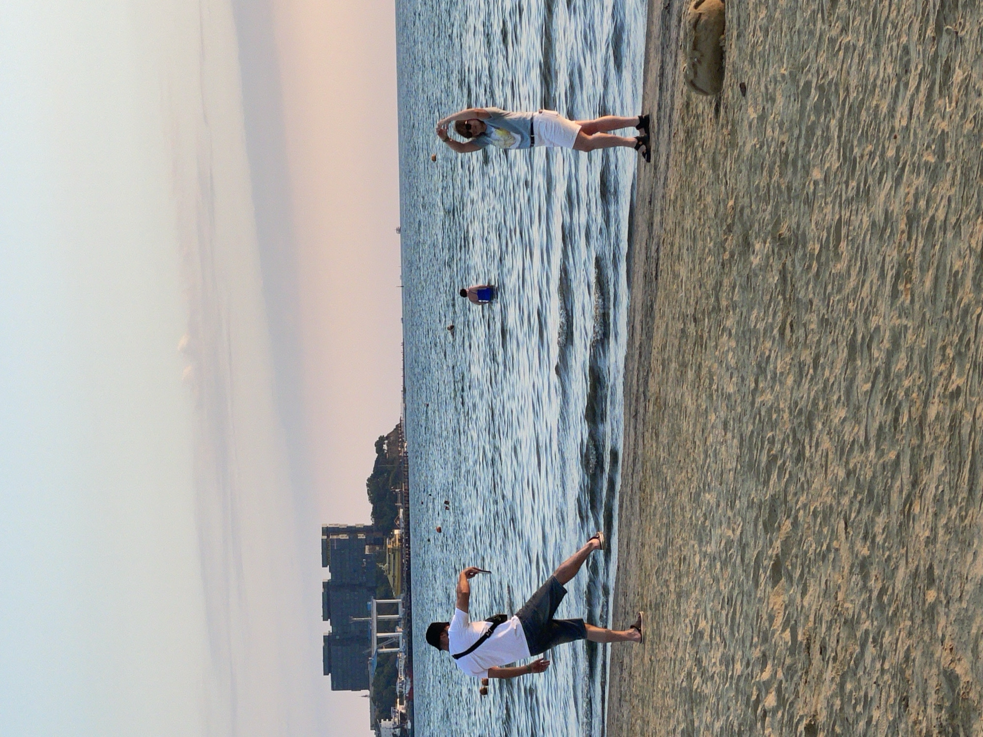 The image depicts a beach scene in Anapa, Russia, with two individuals positioned near the water's edge. On the left, a man wearing a white t-shirt, dark shorts, and sandals is standing on the sand, with his left leg extended forward as if kicking at the water. He is holding a mobile phone in his right hand. On the right, a woman in a light-colored t-shirt and white shorts stands on the sand with her arms raised above her head, forming a shape with her hands. In the background, the sea stretches towards the horizon, with a few people visible in the water. A large, dark building complex is situated on a hill in the distance. The beach is sandy, and the sky is a pale blue with wispy clouds.