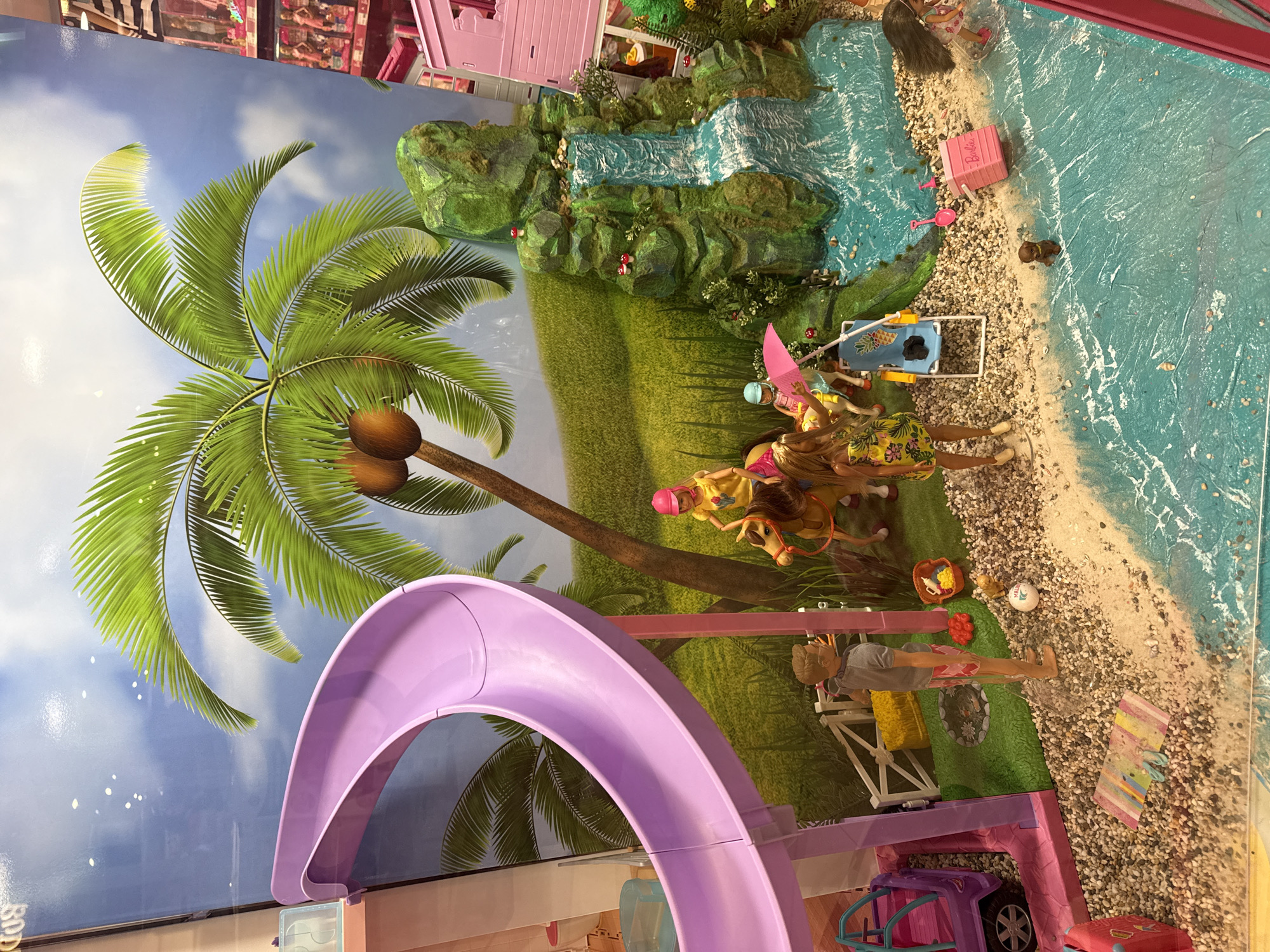 The image displays a Barbie doll playset with a beach and tropical theme. The background features a sky, a palm tree, a waterfall, and grassy terrain. The playset includes Barbie dolls, horses, a slide, a lounge chair, and other accessories. The location in the city of Moscow, Russia, is not discernible from the visual information.