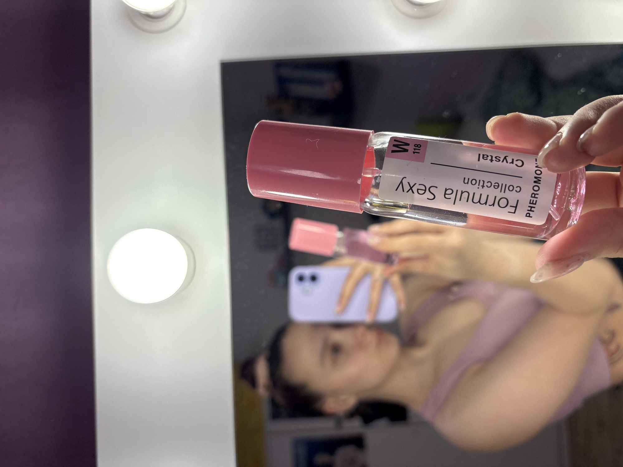 A woman is holding a bottle of "Formula Sexy" pheromone crystal collection perfume in front of a mirror. The mirror is surrounded by lights. The woman is visible in the reflection, wearing a light purple top and holding a purple phone.