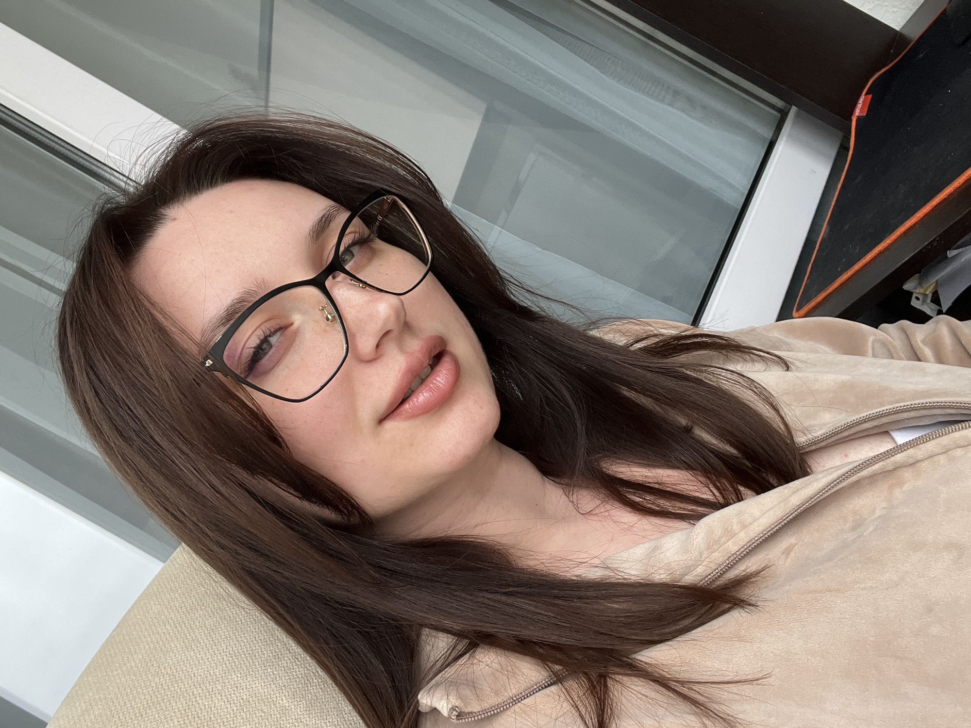 The image presents a close-up portrait of a woman with long, dark brown hair, captured from a slightly elevated and tilted perspective. She wears black-framed eyeglasses with subtle gold accents and has a small stud piercing on her nose. Her lips are softly parted, revealing a light pink, natural shade. She is dressed in a beige or tan zippered jacket, which appears to be made of a soft material like velour or fleece.
The woman is depicted in a reclining or relaxed pose, with her head resting on what appears to be a light-colored, upholstered armchair or couch. Her gaze is directed upwards and slightly to the right, conveying a relaxed or contemplative expression. The lighting in the image suggests it is daytime, with soft, diffused light illuminating her face.
In the background, a large window or balcony door with white frames is visible, suggesting an indoor setting. To the far right, a dark-surfaced desk or shelf is present, along with a black mousepad that has a distinctive orange border. A small red tag on the mousepad clearly displays the text "RAZER". The scene evokes a casual and comfortable atmosphere, likely within a home environment in Tyumen, Russia.