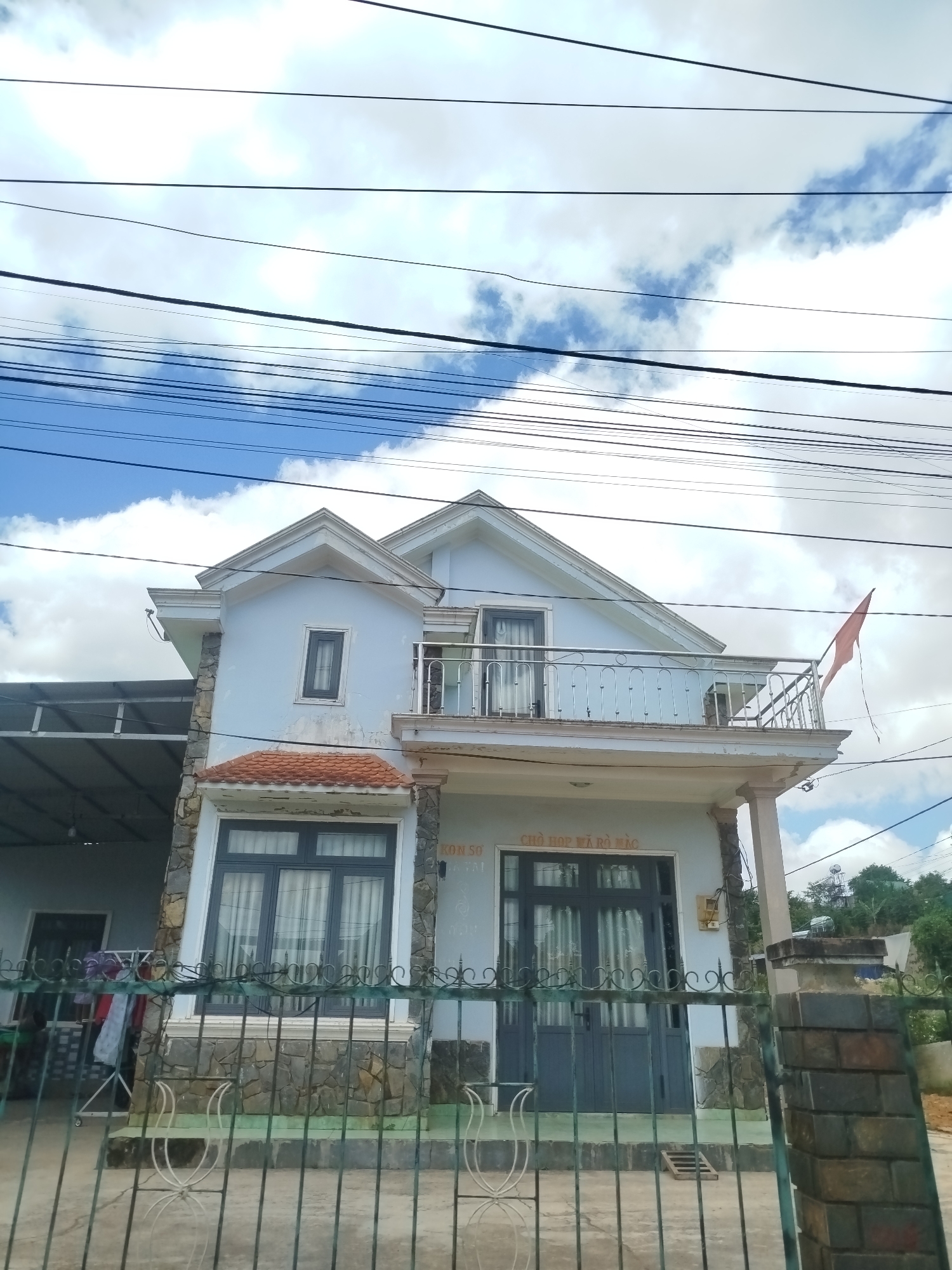 A light blue, two-story house is observed in Thôn 6, Vietnam. The house has a pitched roof with light-colored trim. The exterior walls are a pale blue, with stonework on the lower portions of the house and around a prominent window on the first floor. This window is dark gray with multiple panes and is covered by white curtains. A smaller dark gray window is present on the second story above this window, also with white curtains. There is a balcony on the second story with a silver railing and an orange flag attached to it. A covered carport is visible to the left of the main house. The house is bordered by a dark green metal fence and a brick pillar to the right. The sky above is blue with scattered white clouds. There are multiple power lines running across the top of the image. No people are visible in the image.
