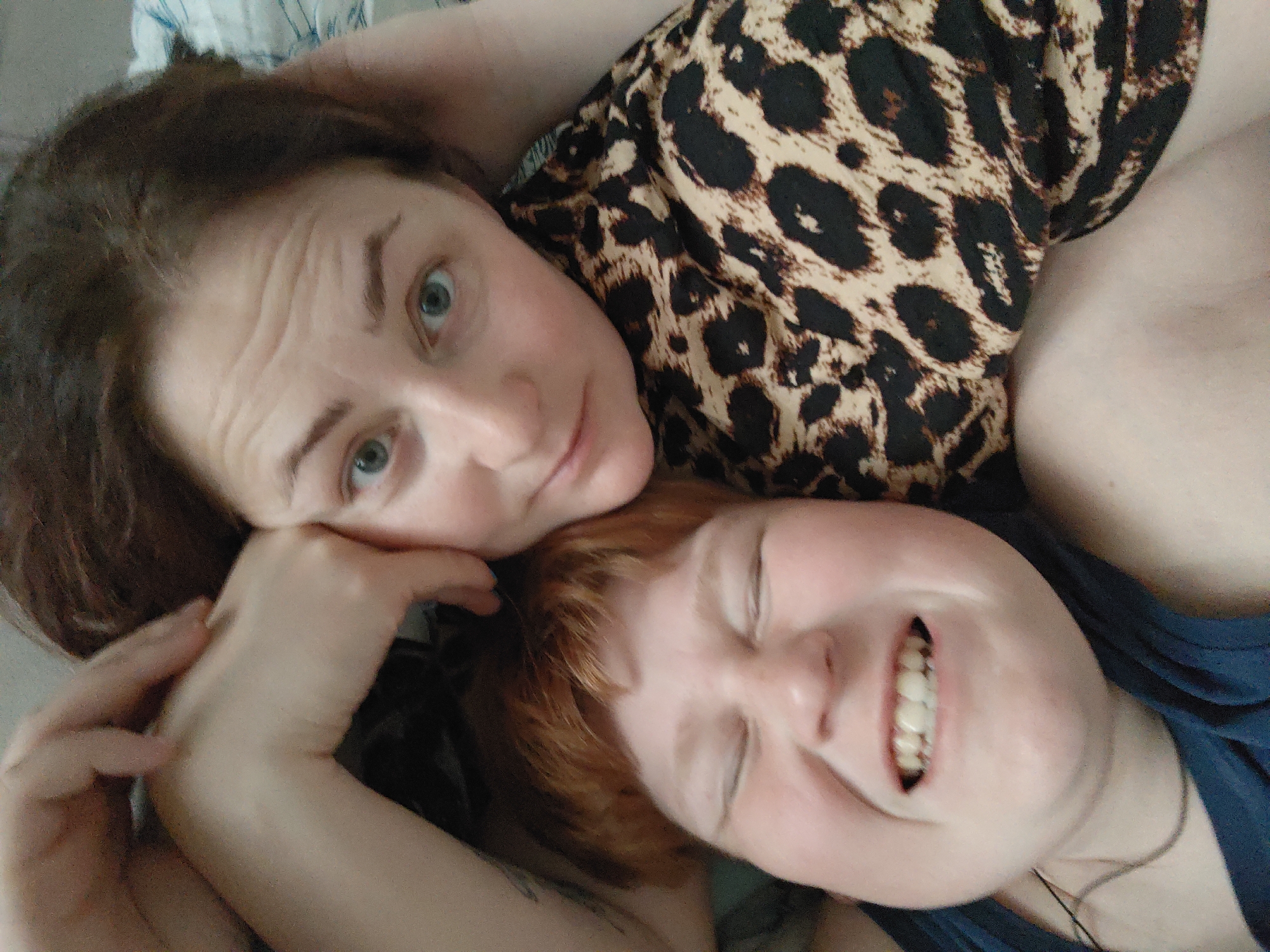 Two women are lying close together. One woman has shoulder-length brown hair and is looking at the camera. She is wearing a leopard print top. The other woman has short red hair and is smiling with her eyes closed. She is wearing a blue top and a necklace. They are lying on a surface with a white and blue pattern. There is no information about the location in the city of Markovskii, Russia.