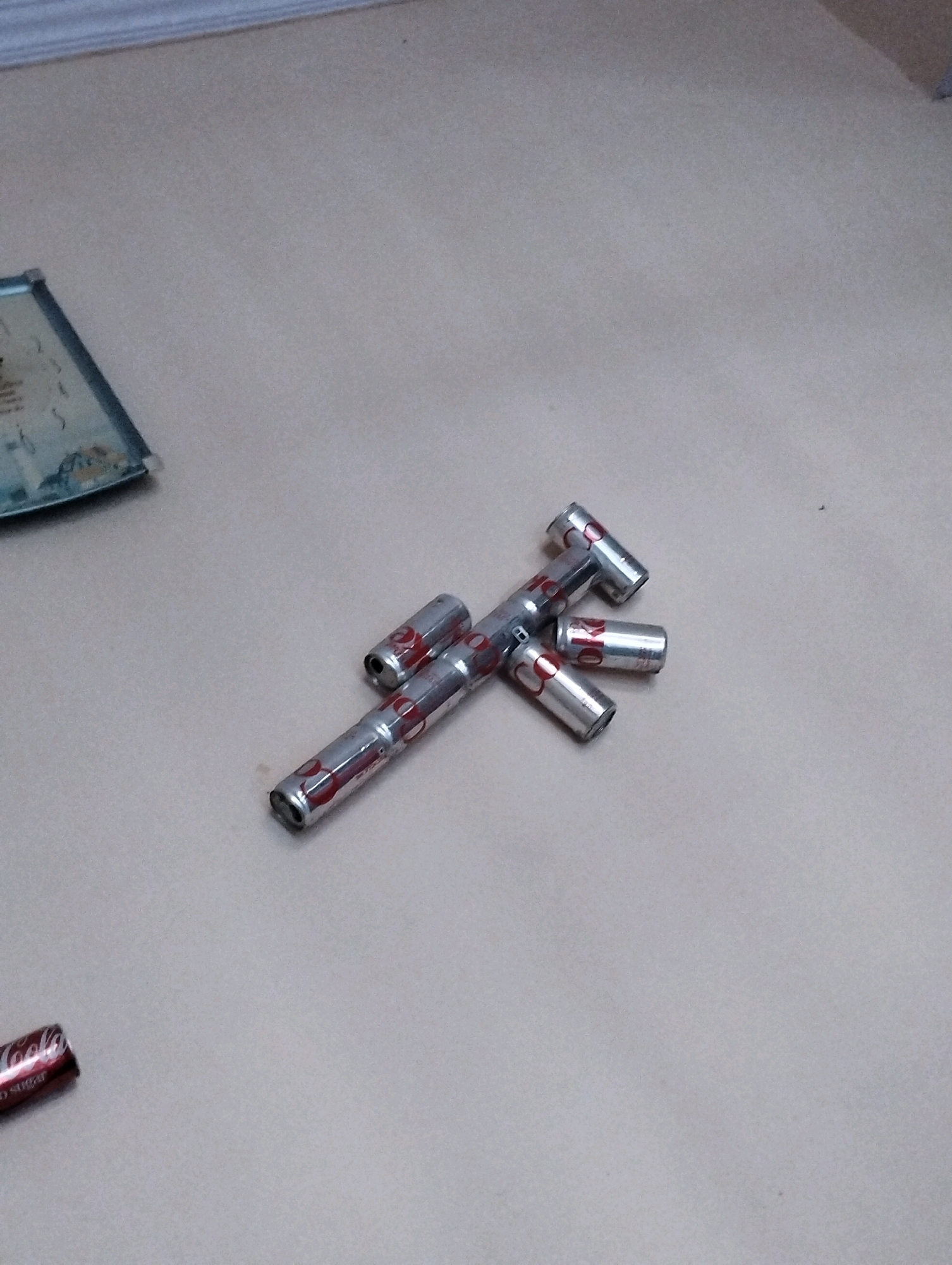 The image displays a close-up, overhead view of several empty, crushed aluminum cans arranged in a cross-like shape on a neutral-colored, textured surface. The cans appear to be from a beverage, with red and silver labeling visible on some. One can at the bottom left of the frame shows red lettering that reads "KOOLa" and "NO SUGAR." The background is a plain, light-colored surface, possibly a table or floor. There is no indication of a city, location, or specific setting within Hathras, India, or any other location. The scene is static and devoid of any human presence or activity.