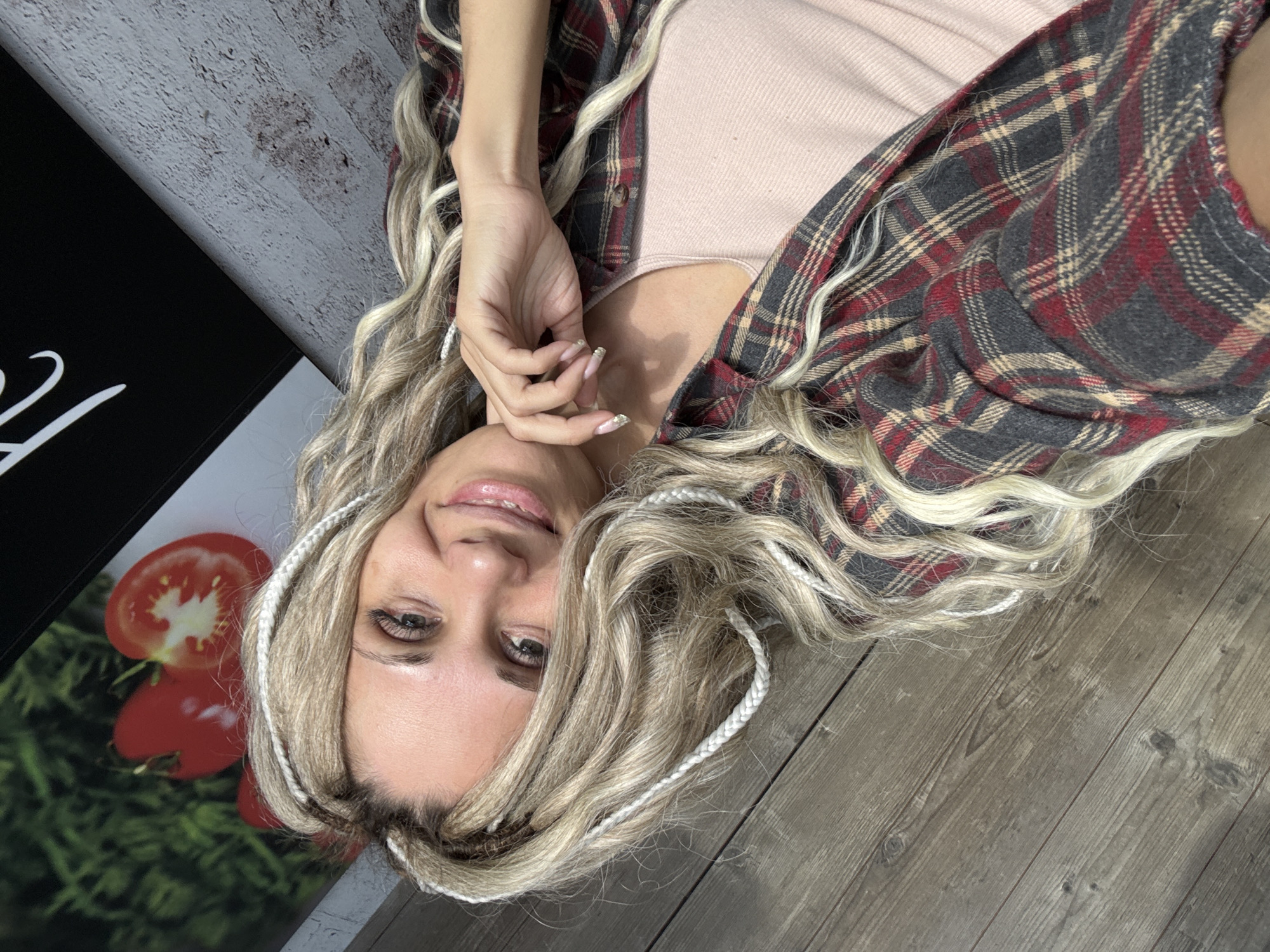 A woman is lying down, facing the camera. She has blonde hair with white braided extensions, and is wearing a pink top and a gray and red flannel shirt. Her hand is near her face. The background includes a black and white poster with a picture of tomatoes, a gray brick wall, and a wooden floor. The image suggests an indoor location, but the location in the city of Rusanovo, Russia is not discernible from the given information.
