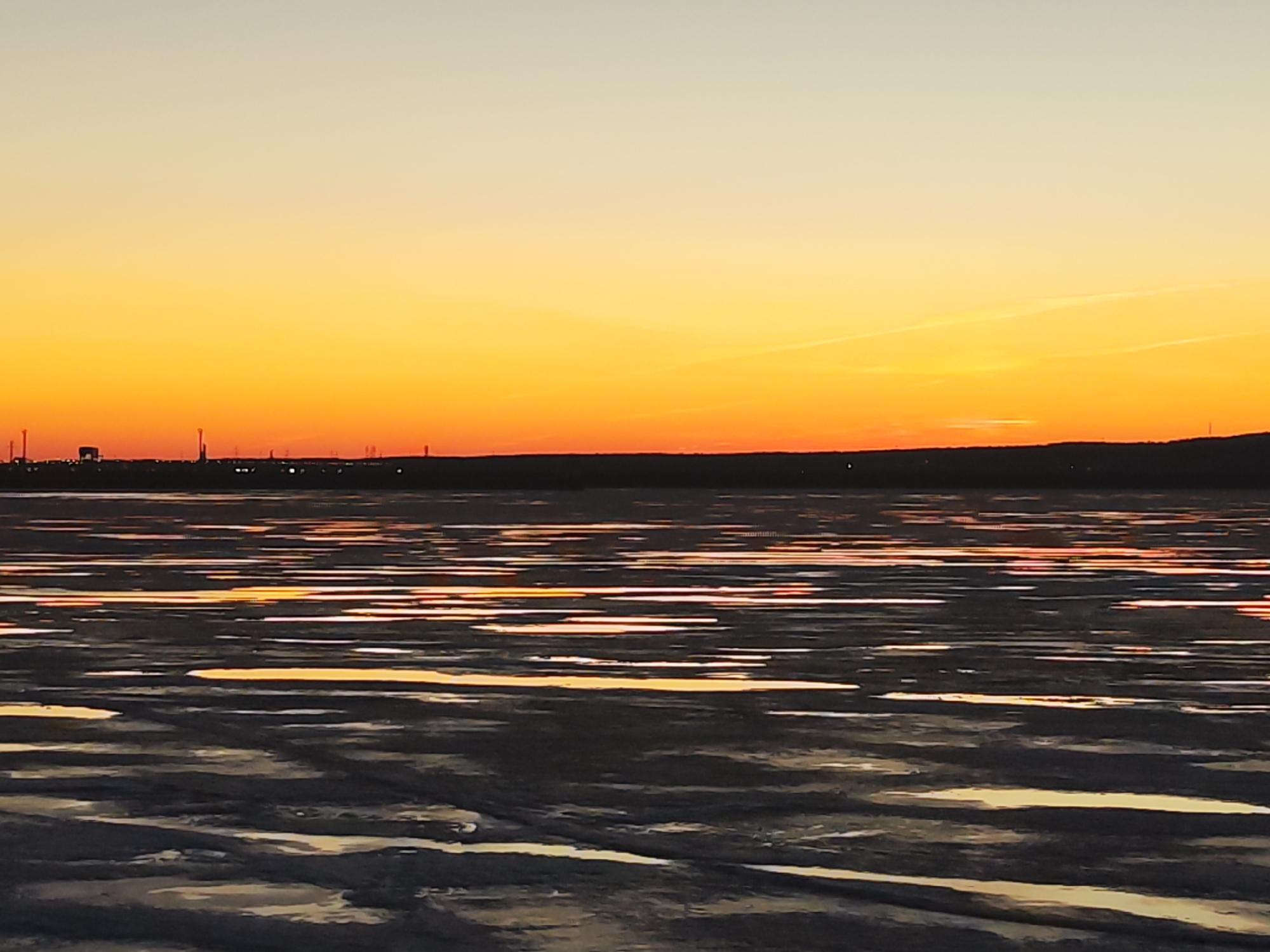 The image shows a sunset over a body of water in Chaikovsky, Russia. The sky transitions from orange to yellow. The water reflects the colors of the sunset. Dark land is visible on the horizon. There are some industrial structures visible on the left side of the horizon line. The surface of the water appears partially frozen.