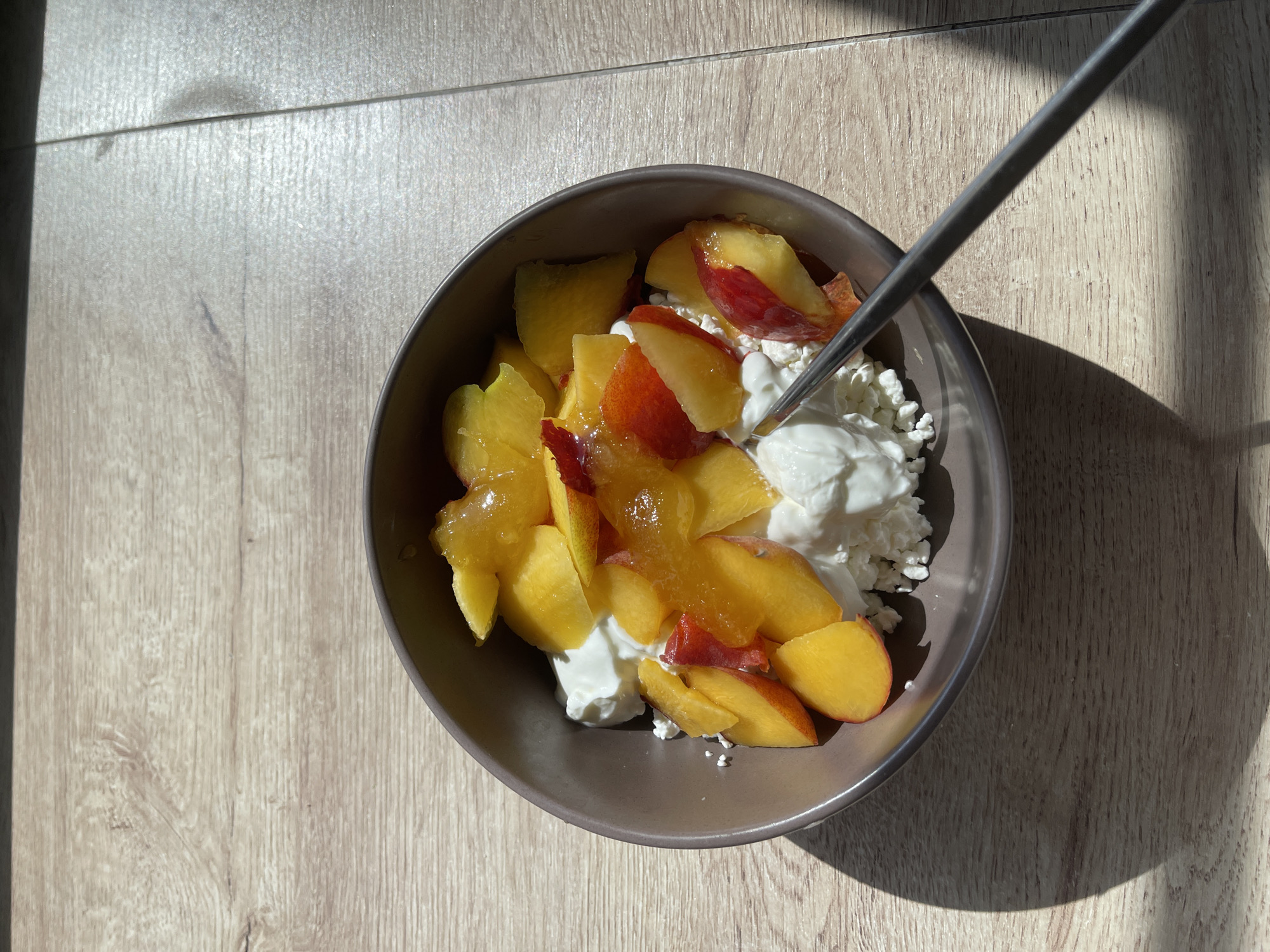 An overhead shot captures a brown-grey bowl filled with a healthy snack on a light wooden surface. The bowl contains white, lumpy cottage cheese or ricotta, topped generously with bright yellow and red sliced peaches or nectarines. A golden, viscous liquid, possibly honey or syrup, is drizzled over the fruit. A slender, metallic utensil, appearing to be a spoon or fork handle, extends into the bowl from the upper right, partially submerged in the white dairy product.
Strong natural light from an unseen window casts distinct shadows across the wooden surface, indicating bright sunshine and suggesting either morning or late afternoon. A dark, elongated shadow from the utensil falls across the bowl and the surface. The wooden surface has a visible grain pattern with horizontal planks. There are no visible people or text in the image. The setting appears to be indoors, possibly at a table or floor near a window, in a location like Мосметаллоконструкция Онт, Russia.