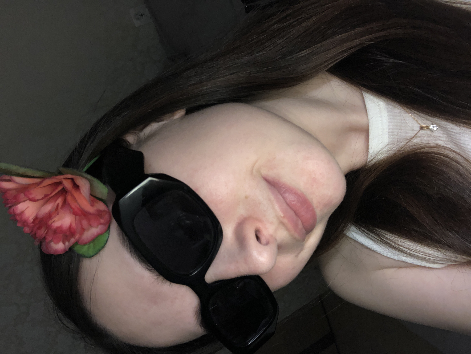 A young woman with long, dark brown hair is positioned with her head tilted toward the left side of the frame. She wears large, black, square-framed sunglasses, and a reddish-pink flower is tucked into her hair near the sunglasses' frame. The woman is wearing a white tank top and a gold necklace with a single, small gemstone pendant is visible around her neck. The background is dark and indistinct.