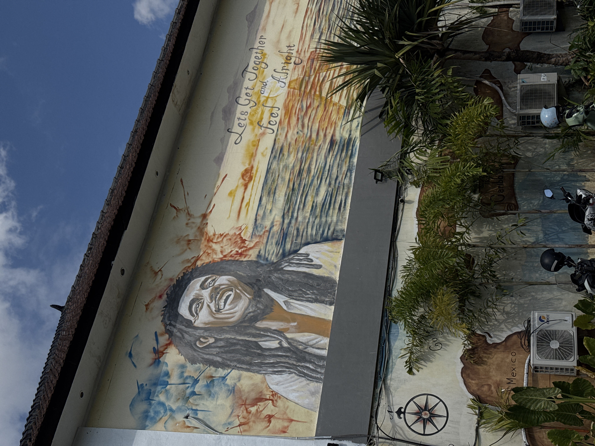 The image depicts a building in North Kuta, Indonesia, adorned with a mural of Bob Marley. The mural also features the phrase "Let's Get Together and Feel Alright" above an abstract landscape, and beneath the portrait there are splatters of blue and red paint. To the right of the mural, there are trees and air conditioning units, and further right are motorbikes, with a map of Mexico and other places in the mural.