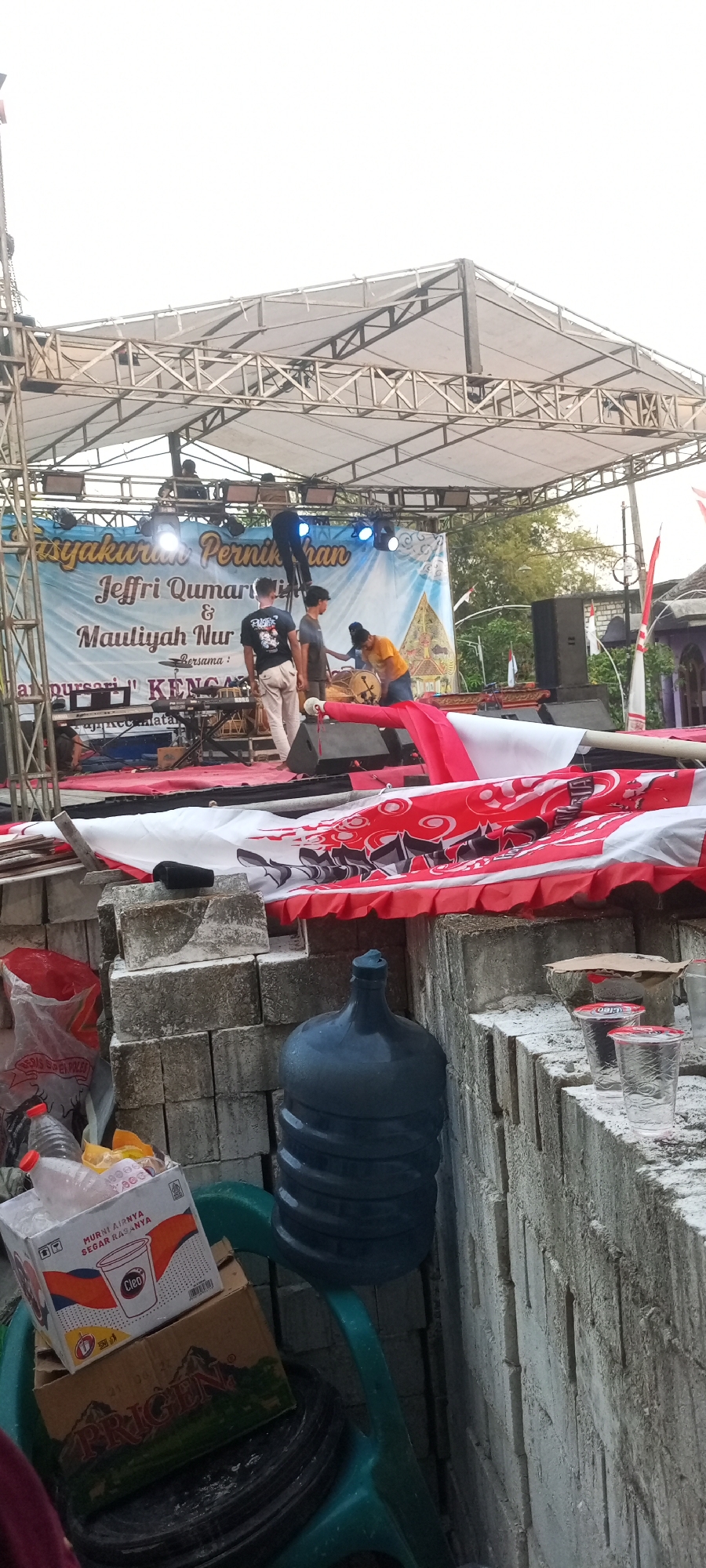 The image depicts an outdoor stage setup in Paji, Indonesia, likely for a community event. The stage is covered by a white canopy supported by a metal truss structure. A banner hangs across the back of the stage with text that appears to be in Indonesian, possibly related to a wedding reception or a similar celebration. Several people are visible on and around the stage. Some individuals are working on the stage lighting, while others appear to be setting up musical instruments. In the foreground, there are stacks of concrete blocks, a large dark blue water jug, and some plastic cups. There are also several boxes of bottled water and other items piled on a green plastic chair. A red and white fabric banner is draped over the front of the stage area. The overall scene suggests a preparation phase for an event.