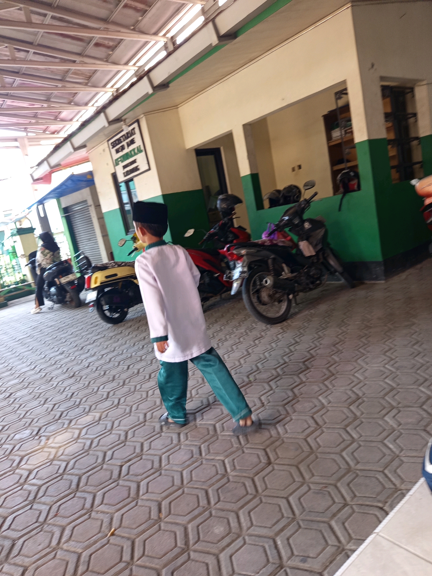 In Karawang, Indonesia, a young boy wearing a white shirt and green pants walks on a patterned tile walkway outside a building. The building has a cream-colored facade with green trim. Several motorcycles are parked in front of the building. In the background, a woman in a hijab is seen near a motorcycle. The area appears to be a public space or educational institution, possibly a school.