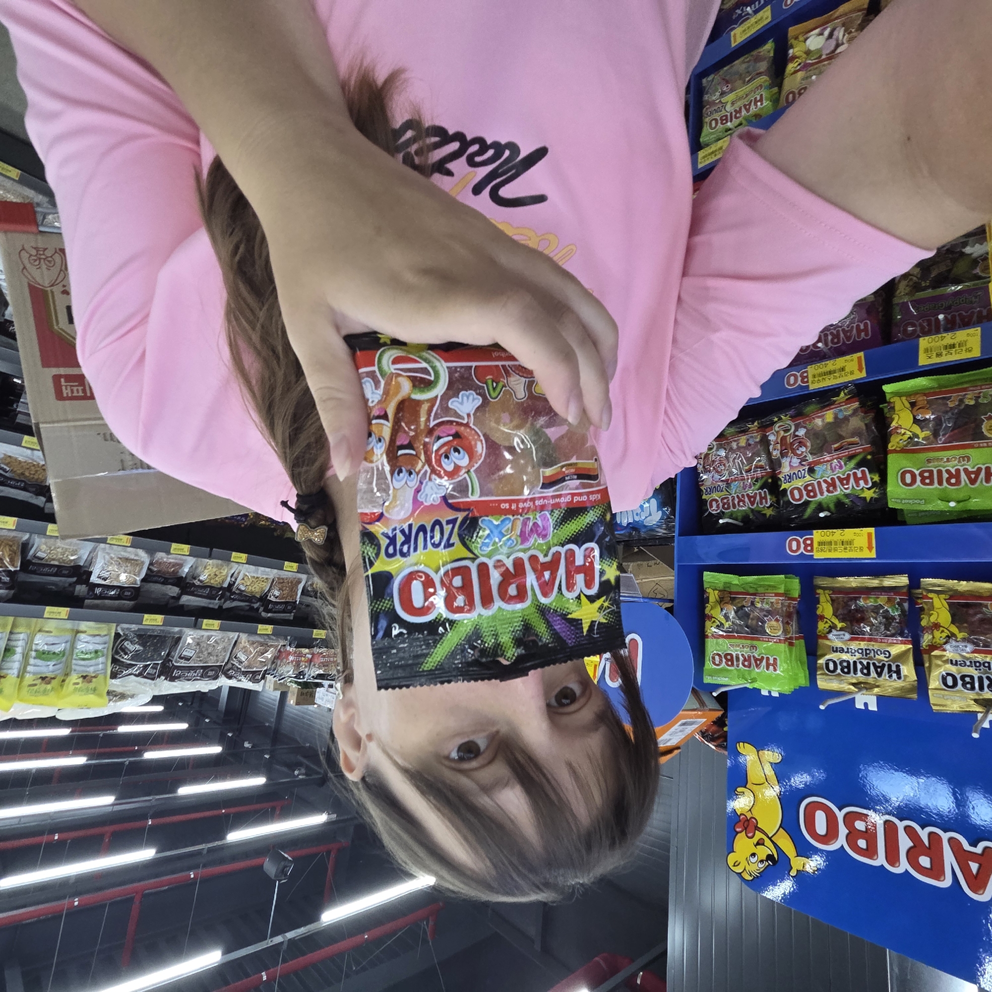 In Dunnae-myeon, South Korea, a person wearing a pink t-shirt is holding a packet of Haribo Sour Zourr gummies in front of their face. The background shows a retail environment with shelves stocked with various Haribo candy products, including Goldbears and other gummy varieties. The lighting appears to be from overhead fluorescent fixtures. The person's face is partially obscured by the candy packet.