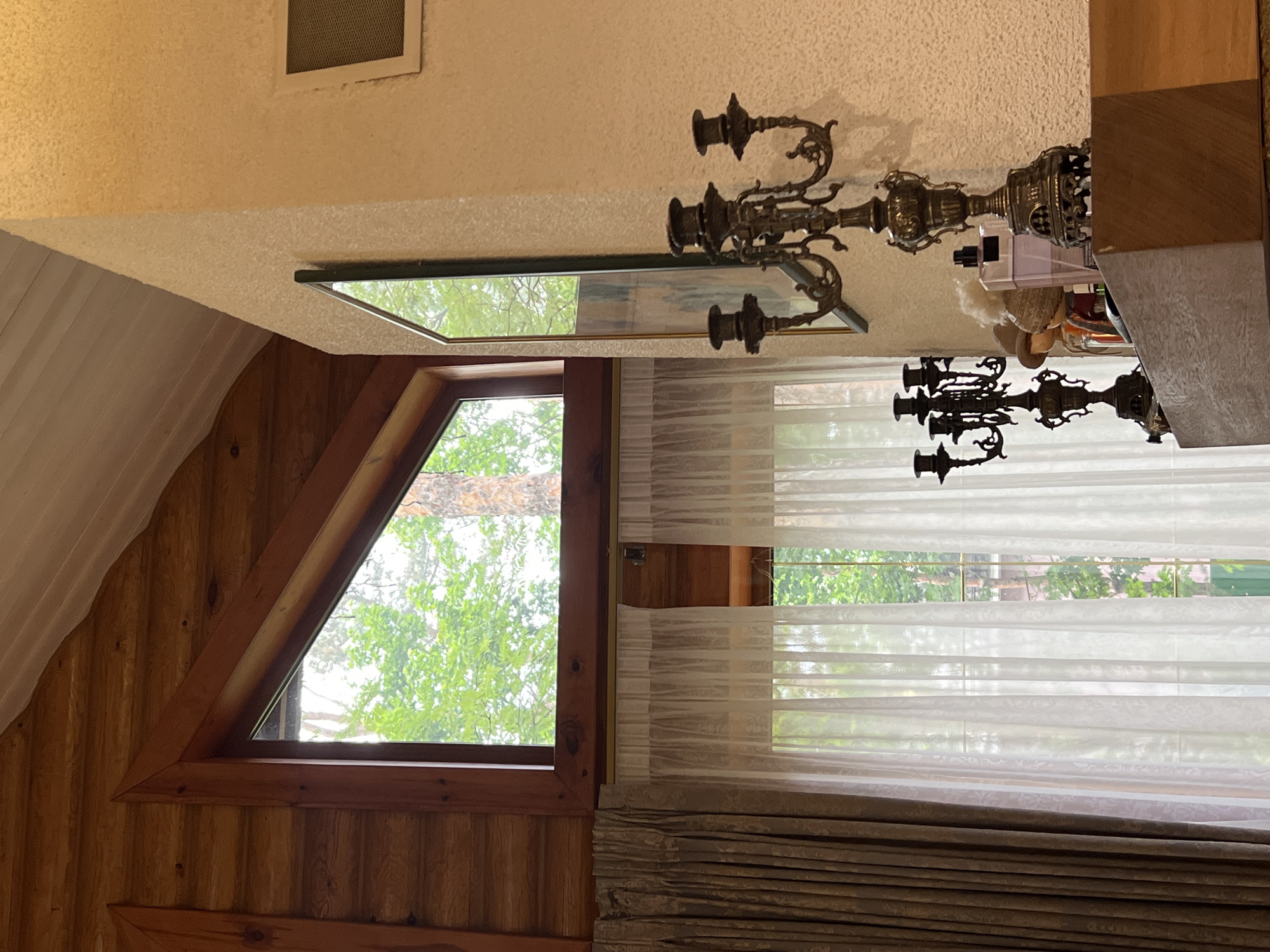 A room in Tomilino, Russia, features a triangular window with a view of green trees. The room has wood-paneled walls on one side. Sheer white curtains are draped over a window beside it, and a gray fabric valance is below. A large ornate, bronze candelabra is mounted on the wall near a mirror. A vent is present in the ceiling.