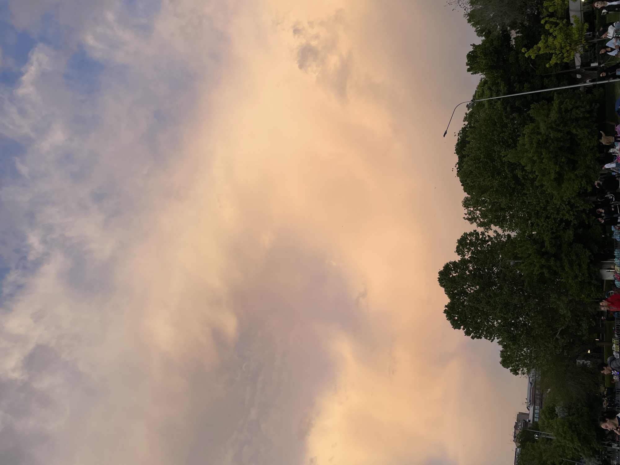 The image captures a sky at dusk with clouds tinted in shades of orange and blue, potentially in Yerevan, Armenia. A row of trees lines the right side of the frame, with hints of buildings visible below. People are present near the tree line. A tall light pole extends from the trees toward the sky.