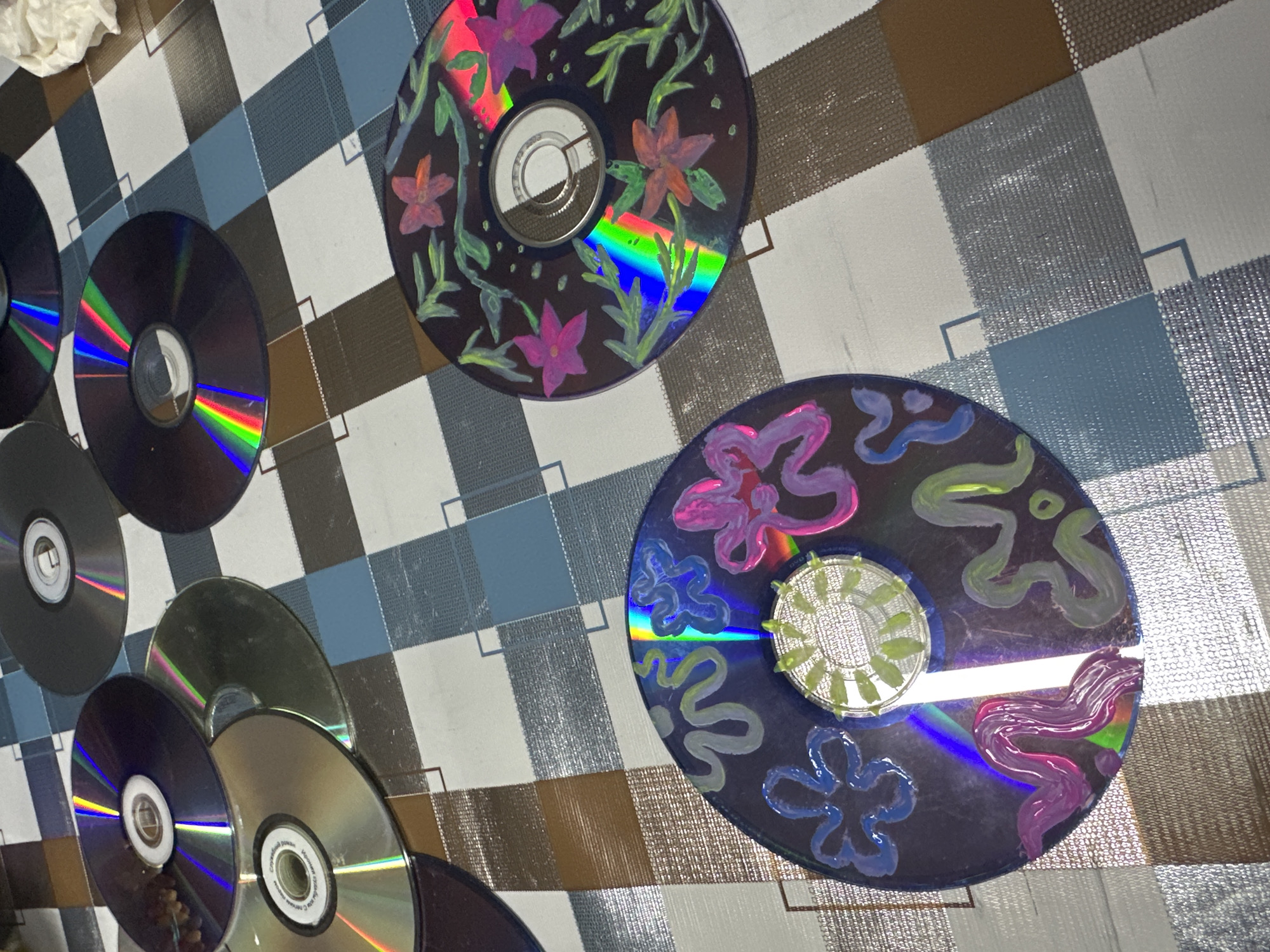 The scene depicts several compact discs (CDs) arranged on a patterned tablecloth. Two of the CDs are decorated with painted floral designs. One features pink and red flowers with green leaves, while the other has blue, pink, and green floral patterns surrounding its center. The remaining CDs are plain and reflect light, showing rainbow patterns. The background is a tablecloth with a checkered pattern of white, blue, and brown squares. A crumpled white object is partially visible in the upper left corner. The location is unknown.