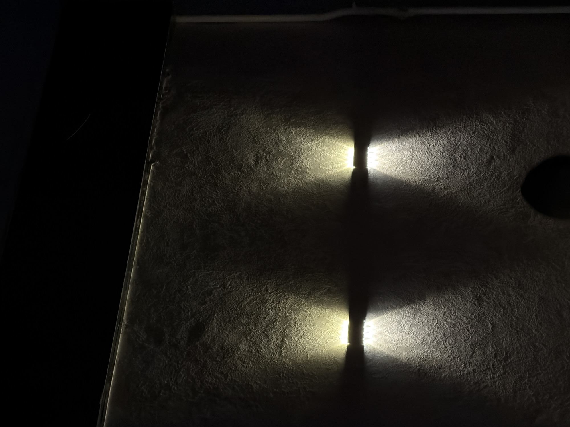 The image shows an outdoor wall illuminated by two downward-facing light fixtures. The wall has a textured, stucco-like finish. The light fixtures are mounted horizontally, and they cast pools of light downwards, creating shadows and highlighting the texture of the wall. The scene appears to be at night, as the background is dark and the only illumination comes from the lights. There is no discernible location information in the image itself, and therefore no reference to Chirchik, Uzbekistan.