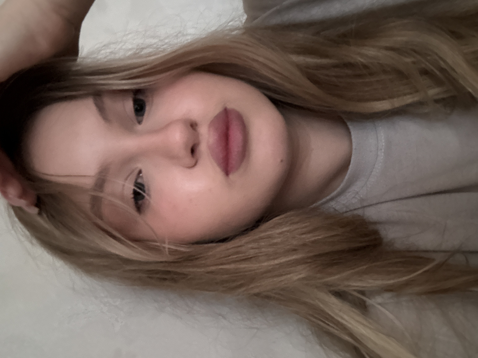 A young woman with blonde hair takes a selfie indoors. She is wearing a grey t-shirt and her left hand is on her head. The background is a plain, textured white wall. The image appears to be taken in a residential setting.