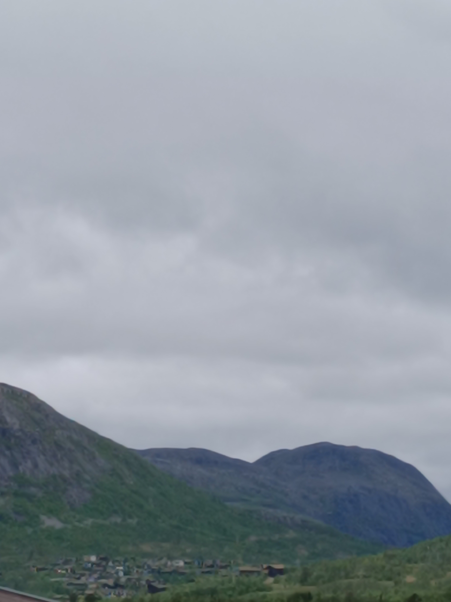 The image depicts a mountainous landscape with a village nestled at the base of the mountains. The sky is overcast with clouds. The village appears to consist of scattered buildings with dark roofs, surrounded by green vegetation. The mountains are rocky and covered with patches of green foliage. The scene suggests a rural setting in a mountainous region, potentially a settlement in Hovden, Norway, given the context. There are no people or animals visible in the image, nor any apparent actions or interactions. The overall impression is one of a quiet, natural environment.