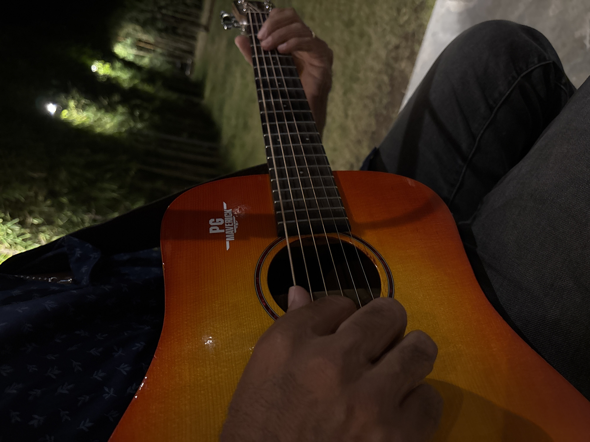 A close-up shot captures a person playing an acoustic guitar. The camera focuses on the guitar's body, which has a sunburst finish in shades of orange and yellow. The person's hands are visible, one strumming the strings near the soundhole and the other fretting on the neck. The guitar has a "PG MAVERICK" logo printed on its body. The setting appears to be outdoors at night, with soft, diffused lighting creating a warm ambiance. In the blurred background, hints of greenery and possibly artificial lights can be seen, suggesting a garden or outdoor seating area. The person is wearing dark jeans, and the overall mood is relaxed and intimate, possibly indicating a casual jam session or a moment of personal enjoyment.