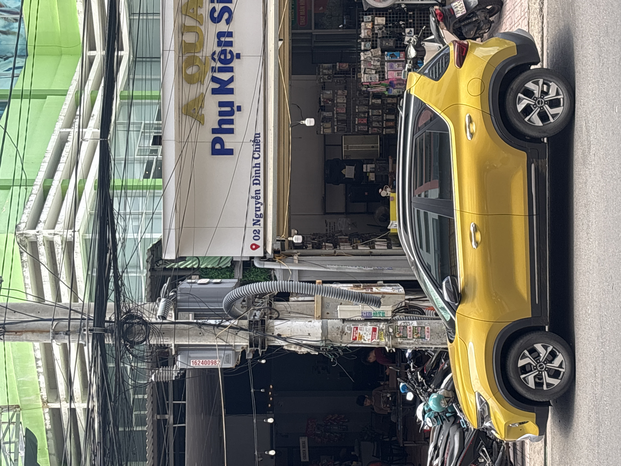 A yellow compact SUV is parked on a paved street in front of a shop with a blue sign that reads "Phu Kien Si." The shop displays various items for sale. Behind the shop, a building with a green facade is visible. A utility pole covered in wires stands next to the shop. Several motorcycles are parked to the left of the SUV. Two individuals are seated inside a shaded area to the left of the shop. The sky is overcast.