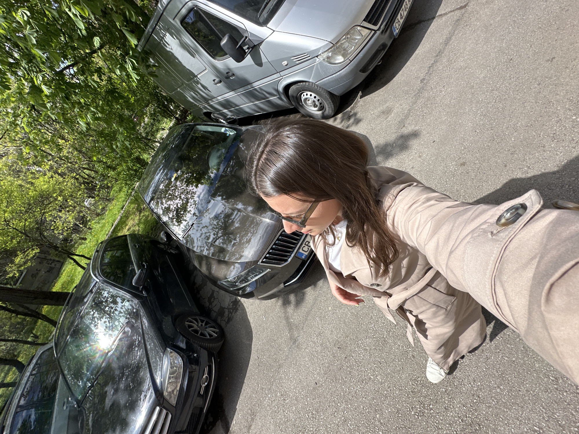 An overhead, slightly tilted selfie captures a woman standing on an asphalt road between parked cars on a sunny day in Sofia, Bulgaria. The woman, with long brown hair, wears glasses, a white top, and a light beige trench coat, holding the camera extended with her left arm. She is looking down and to her right.
To her left, a dark-colored sedan is parked, partially obscured by the woman. Its front grille and a partial "CB" license plate are visible. To her right and behind, a silver Mercedes-Benz Sprinter van is parked, with its full license plate, "CB 4748 KH," clearly visible. "CB" indicates Sofia, confirming the provided location context.
The setting appears to be an urban or suburban street with ample green foliage and trees along the sides, suggesting a leafy area or park nearby. Bright sunlight casts distinct shadows on the asphalt, indicating clear weather. The overall impression is a casual, bright outdoor scene.