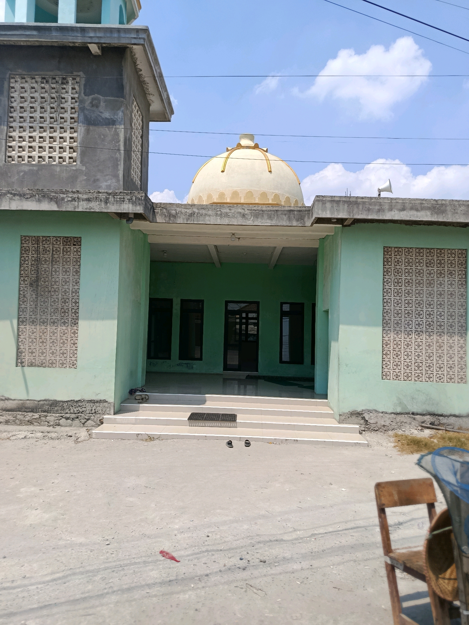 The image shows a mosque in Kedungpengawas, Indonesia. The mosque has a light green facade and a cream-colored dome. The entrance to the mosque is a large open doorway with steps leading up to it. There are several windows on the facade of the mosque, each with a decorative lattice pattern. The sky above is blue with some white clouds. In the foreground, there is a dirt road with some debris on it, including a piece of red plastic and a pair of sandals. There is also a wooden chair and what appears to be a fishing net or basket in the lower right corner of the image. The overall scene depicts a sunny day in a rural or semi-rural setting.