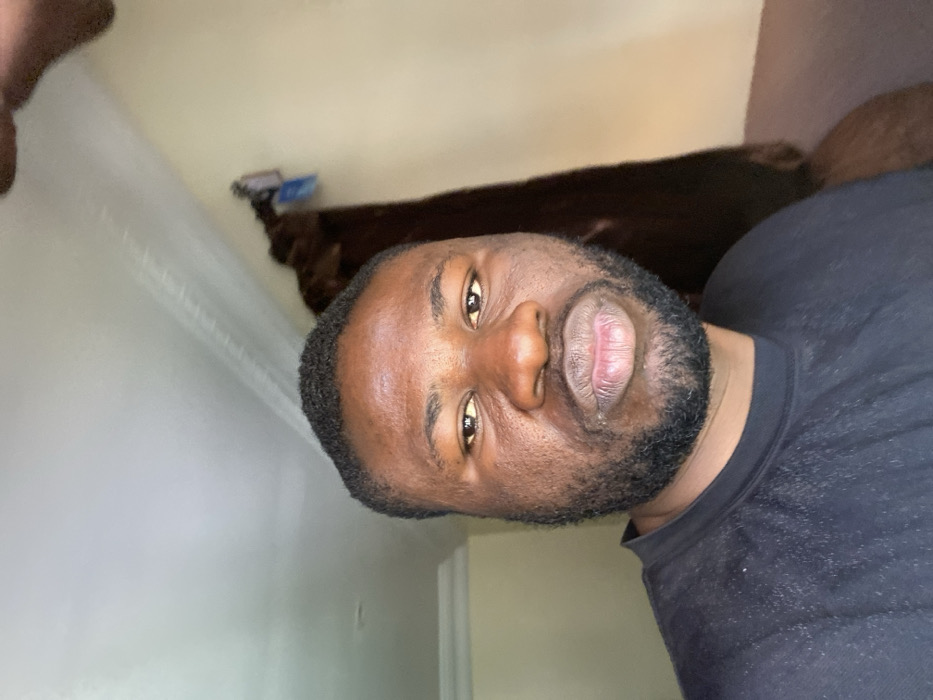 The scene depicts an indoor environment in Udu, Nigeria. A single adult male is featured in a head-and-shoulders shot, occupying the central and left portions of the frame. The individual has dark skin, short dark curly hair, a full beard, and a mustache. He wears a plain black crew-neck t-shirt or sweatshirt, with minor light-colored particulate matter visible on the fabric of his left shoulder and chest. His eyes are dark, and he maintains a direct gaze towards the camera. His lips are closed and appear slightly pursed. The background consists of a light-colored wall, possibly cream or light yellow, with white crown molding visible along the top left edge. To the right of the subject, a dark, vertical object, consistent with a curtain or heavy fabric, is partially visible. A small, light-colored item is hanging from the upper right portion of this dark object.