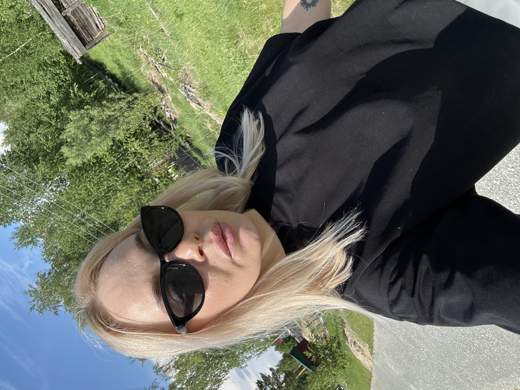 In this frontal, eye-level selfie, a blonde woman wearing black sunglasses and a black t-shirt poses outdoors. Her lips are slightly parted, and her hair is loose and falls around her shoulders. The background features a bright blue sky with scattered clouds, lush green trees, and a rustic wooden structure that appears to be a shed or small building. The ground is covered in grass and a dirt path. A small tattoo of a sun-like design is visible on the woman's right arm. The scene suggests a rural or natural setting.
