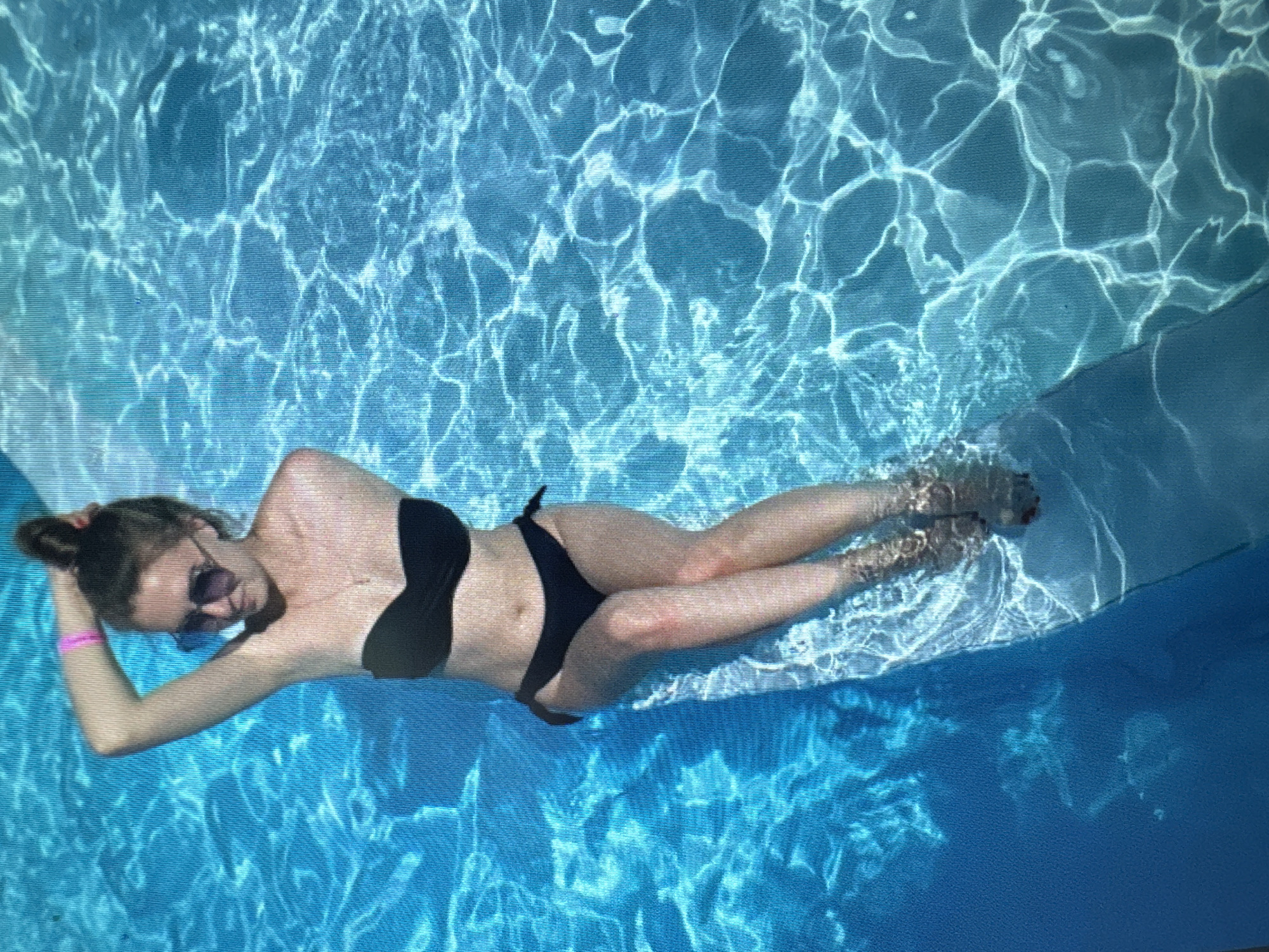 A young Caucasian woman with brown hair pulled into a bun is floating in a swimming pool. She is wearing a black bikini, sunglasses, and a pink bracelet on her left arm. Her left hand is behind her head. The water is bright blue and has ripples and light reflections on the surface. The bottom of the pool is visible and appears to be tiled. The woman is positioned diagonally in the frame, with her head towards the top left and her feet towards the bottom right.
