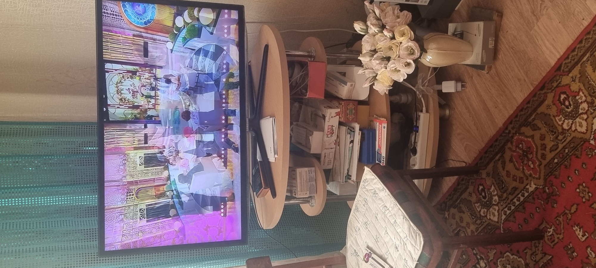 The media file shows an indoor scene with a television displaying an animated image of people in a brightly lit, ornate setting. A wooden table with rounded edges stands beside the television, holding various objects including books, boxes, and a power strip. A bouquet of light-colored flowers sits on top of the table, next to a decorative vase. Further back, a chair with a patterned cushion is visible, and a portion of a red carpet with intricate designs covers the floor. The lighting suggests it is daytime. Some visible text on boxes includes "ГЛАВ ТОРТ" and "ВЕНОЛТОН 911". The overall impression is of a lived-in domestic space, possibly a living room or study. The location context is Krymsk, Russia.