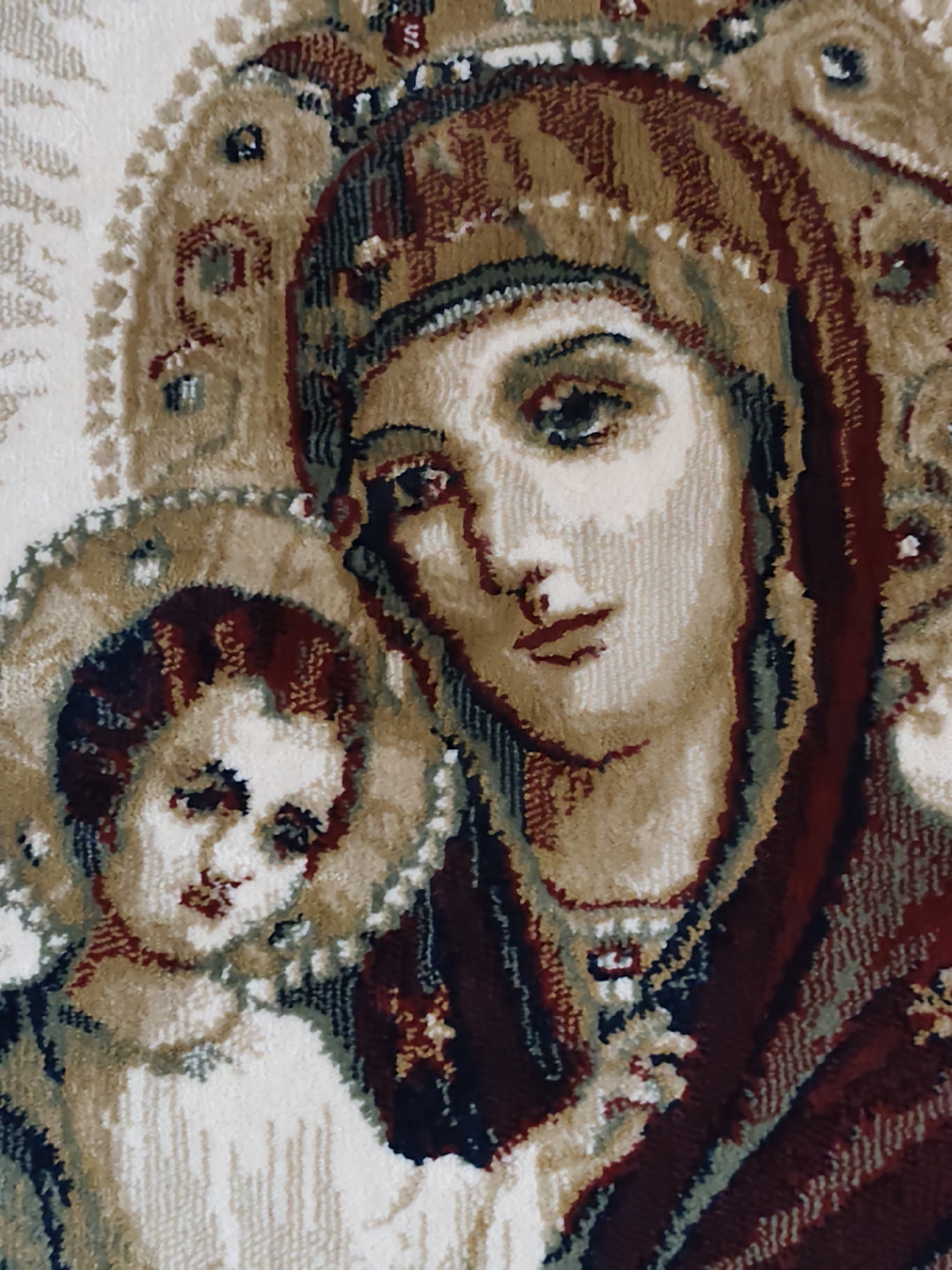 The image is a close-up, textured depiction of a religious icon, specifically the Virgin Mary holding the Christ child. The figures are rendered in a tapestry-like style, with visible threads creating a soft, woven effect. The Virgin Mary's face is central, with expressive eyes and parted lips. She wears a deep red garment and a headdress. The Christ child is positioned to her left, also with visible features and a halo-like circle surrounding his head. The background features decorative, stylized patterns in beige and cream tones. This artwork is located in the city of Ungheni, Moldova.