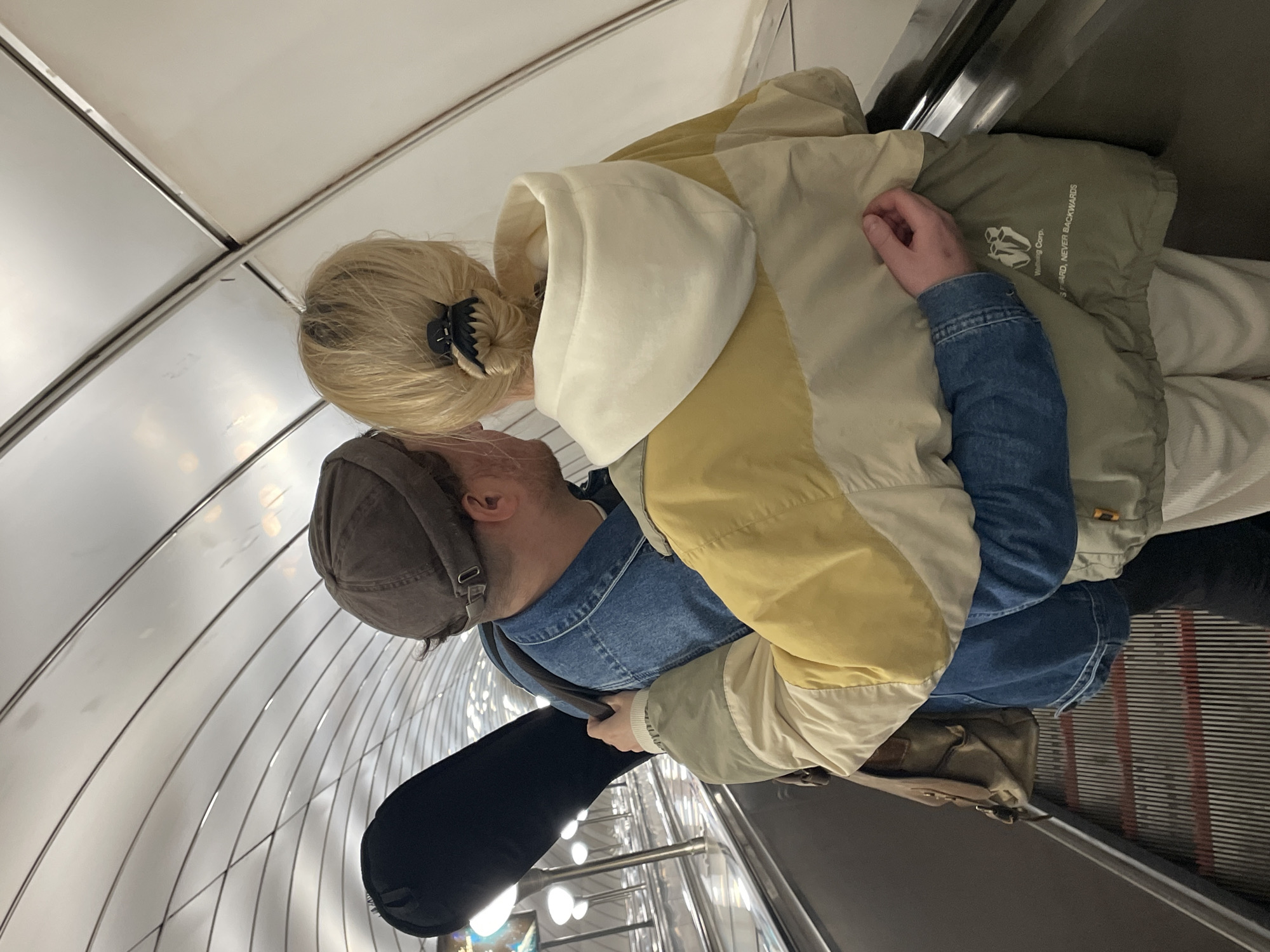 Two individuals are on a subway escalator in Saint Petersburg, Russia. The person in front, with blonde hair pulled into a bun secured by a black clip, wears a beige and yellow color-blocked jacket over a white hooded sweatshirt. The person behind them, wearing a brown cap and a denim jacket, has their arm around the front person's waist and is carrying a black case, possibly for a musical instrument, over their left shoulder. Their faces are obscured as they lean towards each other, suggesting an intimate moment. The escalator is enclosed by a curved, white ceiling with metallic accents and features overhead lighting. The steps of the escalator are metallic and striped with red.