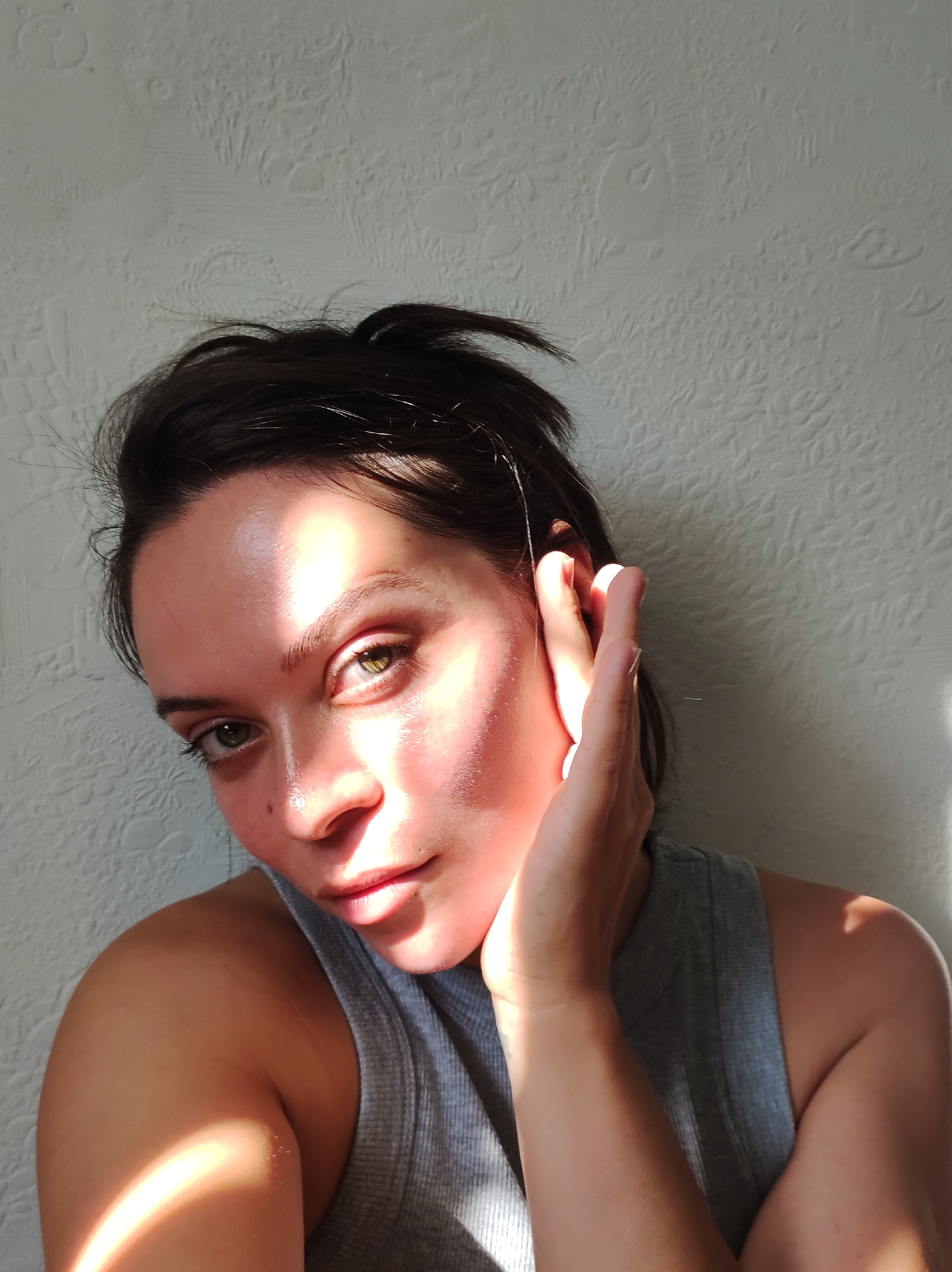 A close-up, high-angle shot shows a person's face illuminated by strong sunlight, creating a halo effect around their head and casting shadows. The person has dark hair styled loosely, with strands falling across their forehead. Their right hand is raised to their ear, obscuring part of their face. They are wearing a gray tank top. The background is a textured white wall. The image was captured in Chaikovsky, Russia, based on metadata.