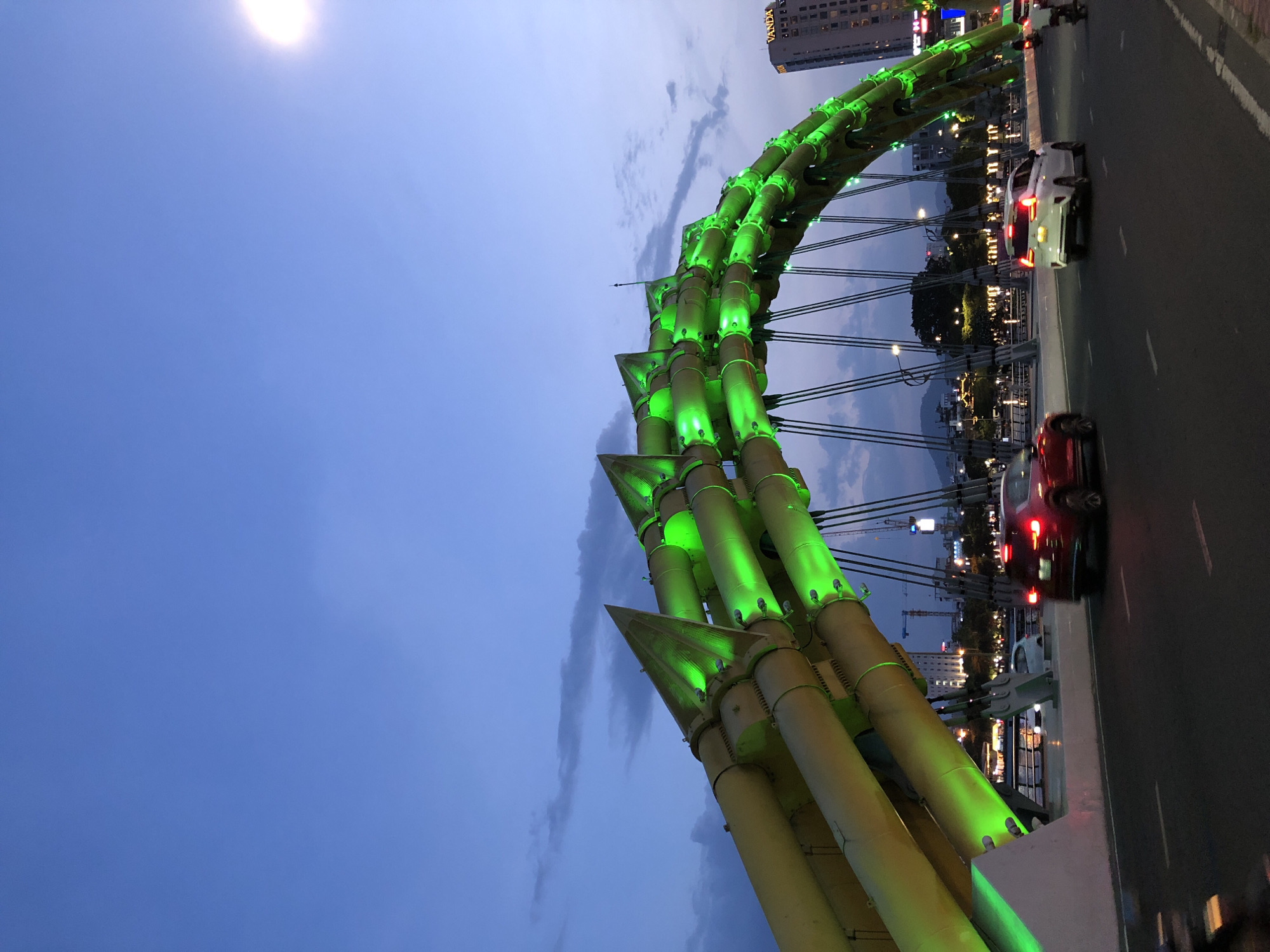 The image shows the Dragon Bridge in Da Nang, Vietnam, at dusk. The bridge is illuminated with green lights, and its distinctive dragon-like structure is visible. Cars are driving on the bridge, and there are buildings and trees in the background. The sky is a deep blue with some clouds, and the sun is setting on the right side of the image. The overall scene depicts a modern urban landscape with a prominent architectural landmark.