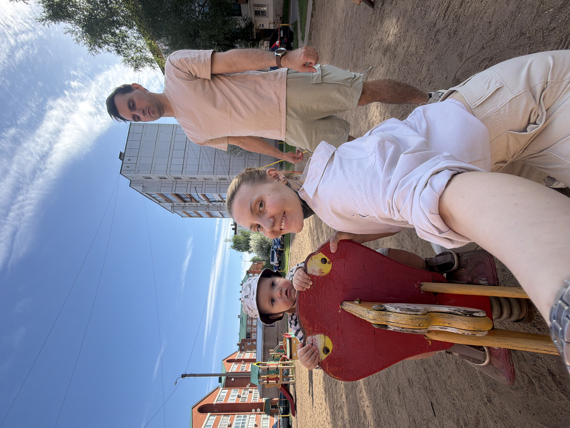 A family of three is in an outdoor playground. The mother, in a white shirt, is holding the camera and taking a selfie. She is positioned near a red and yellow wooden spring rider, where her baby sits in a striped shirt and hat. The father, in a pink shirt and beige shorts, is standing behind and to the side of the mother.
The playground is located in a sandy area, with other playground equipment visible in the background. In the distance, there are multi-story buildings, including a red brick building with white-framed windows. The sky is blue with scattered white clouds.
Based on the visible architecture and playground equipment, this photo appears to have been taken in Chaikovsky, Russia.