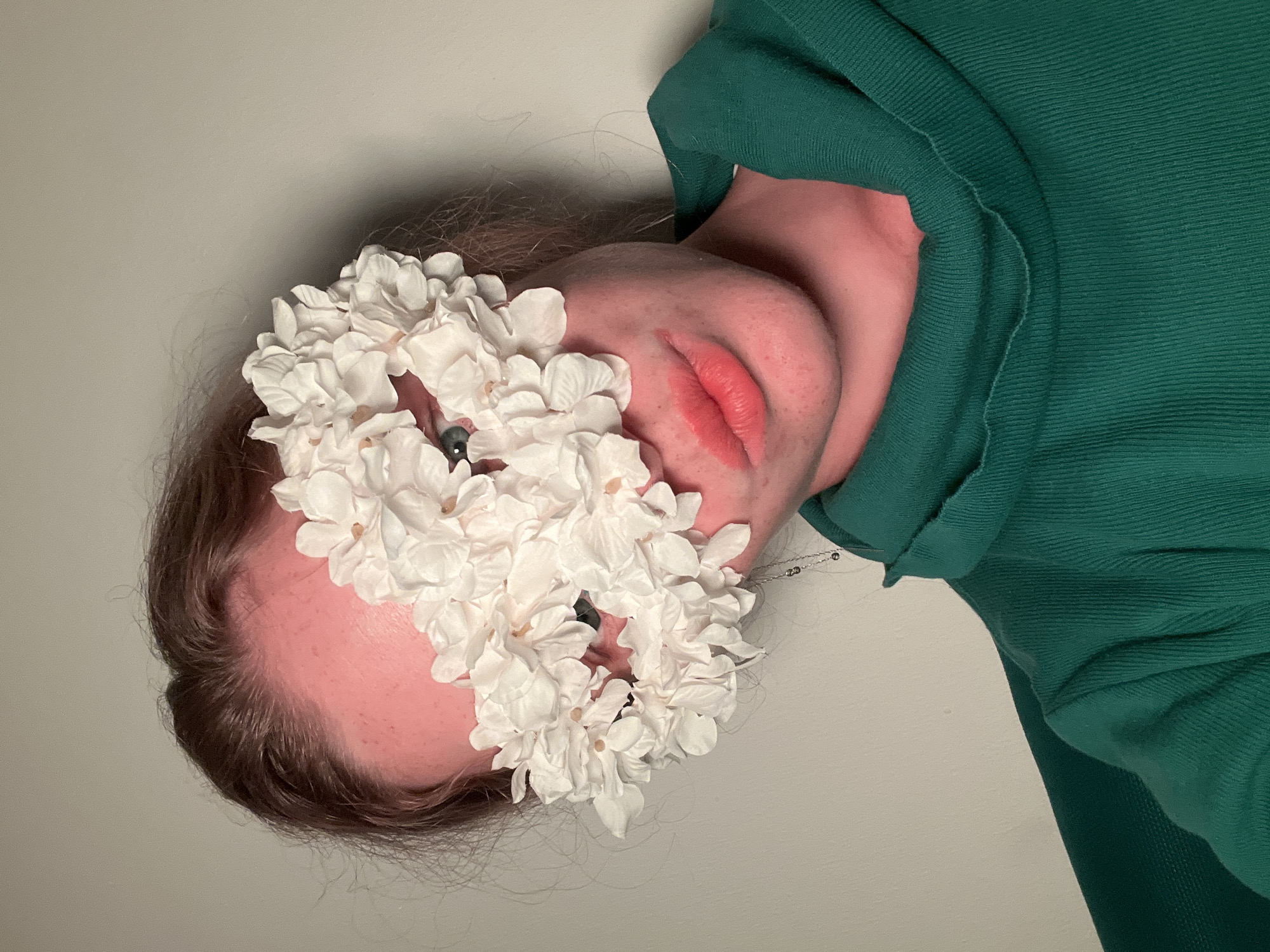 A close-up, rotated image displays a person's face partially obscured by a cluster of white flowers. The person is wearing a green, textured, hooded sweatshirt. Visible facial features include a portion of their forehead, an eye, and lips with visible lip color. The background is a plain, light-colored wall. There is no indication of the image's location being in Budva, Montenegro.