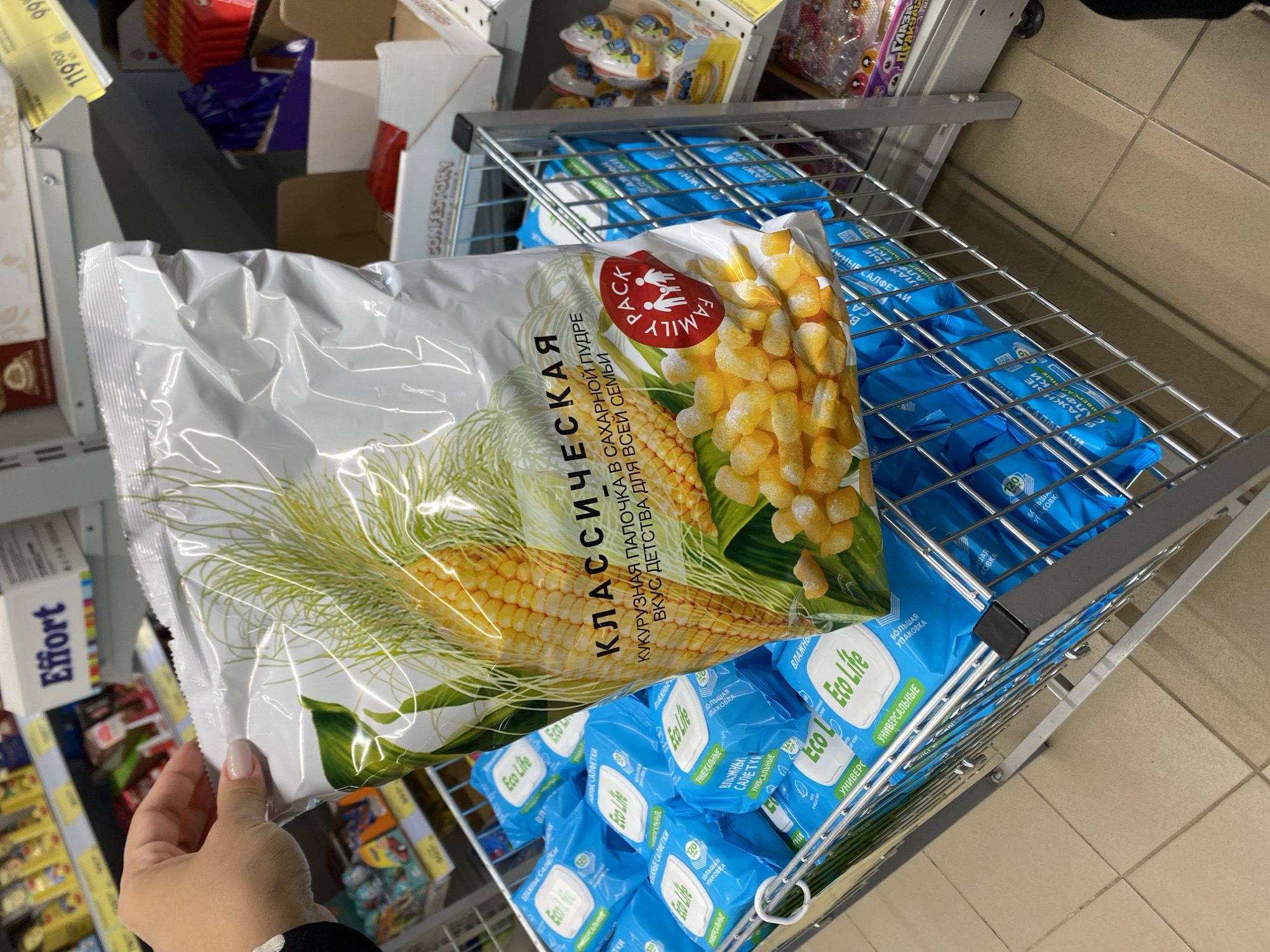 A person's hand with light-colored nail polish is holding a large, clear plastic bag of corn snacks. The bag features an illustration of a corn cob and kernels, along with Russian text that reads "КЛАССИЧЕСКАЯ КУКУРУЗНАЯ ПАЛОЧКА В САХАРНОЙ ПУДРЕ ВКУС ДЕТСТВА ДЛЯ ВСЕЙ СЕМЬИ" and a red "FAMILY PACK" logo with figures of a family. The bag is positioned in front of a shopping cart filled with blue packages of "Eco Life" wet wipes. The shopping cart is in a store aisle, with shelves of products visible in the background. A sign reading "Effort" is visible on an upper shelf. The flooring appears to be tiled. The image was taken in Kushnarenkovo, Russia.