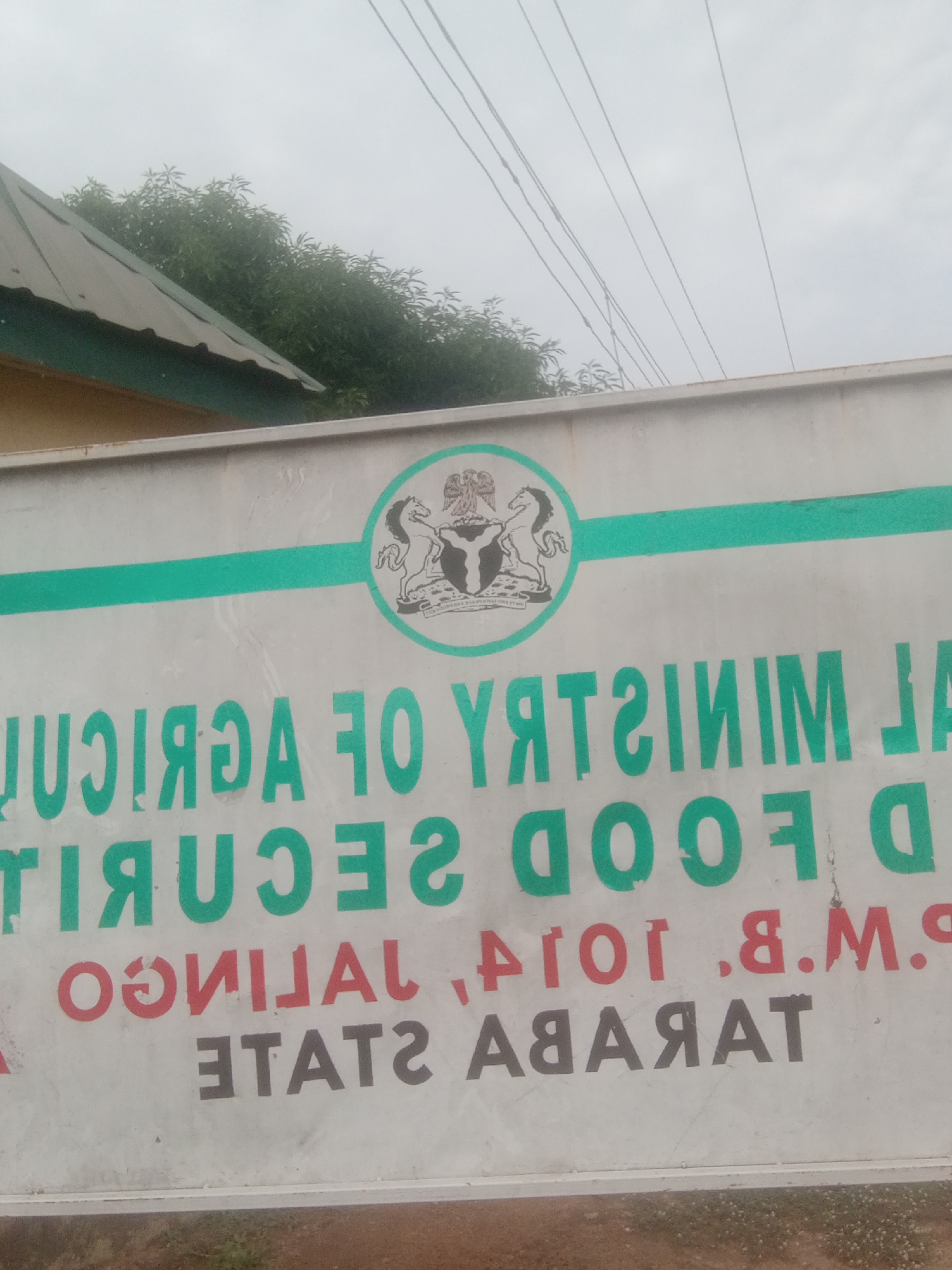This is a close-up, eye-level shot of a sign for the "FEDERAL MINISTRY OF AGRICULTURE AND FOOD SECURITY". The sign is white with green and red lettering. The top part of the sign features the Nigerian coat of arms, a seal depicting an eagle perched on a shield with a wreath. A thin green line runs horizontally across the sign, just below the coat of arms. The text on the sign is in a large, bold font, and appears to be reversed or reflected.
The visible text reads:
"JUQIAƎA ƎO YЯTƎIMIM JA"
"ƧƎCITIᑫꟘ ƧꟘOOꟘ D"
"OƧUIVAL .B.M.9"
"ƎTATE ƧTATE"
The last line, "ƎTATE ƧTATE", is likely meant to be "TARABA STATE", a state in northeastern Nigeria. The context "Jalingo, Nigeria" indicates that Jalingo is the capital of Taraba State. The sign itself is somewhat weathered, suggesting it has been exposed to the elements.
In the background, a portion of a building's roof with a green trim is visible on the left, along with lush green trees. Overhead, several dark electrical wires stretch across the grey, overcast sky. The overall impression is of an outdoor scene, likely during the day, with overcast weather conditions. There are no people visible in the image.
